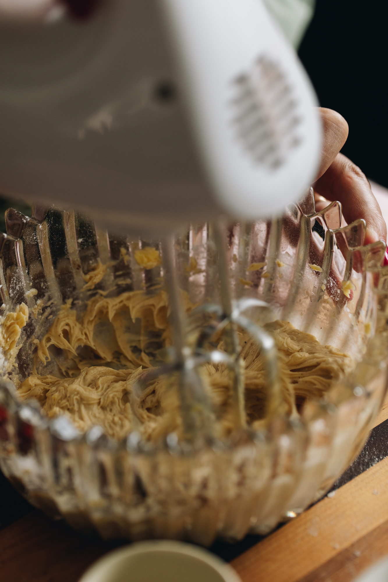 On a striped wooden board sits a glass vintage bowl that has butter in it that is being whipped with a white hand mixer. 