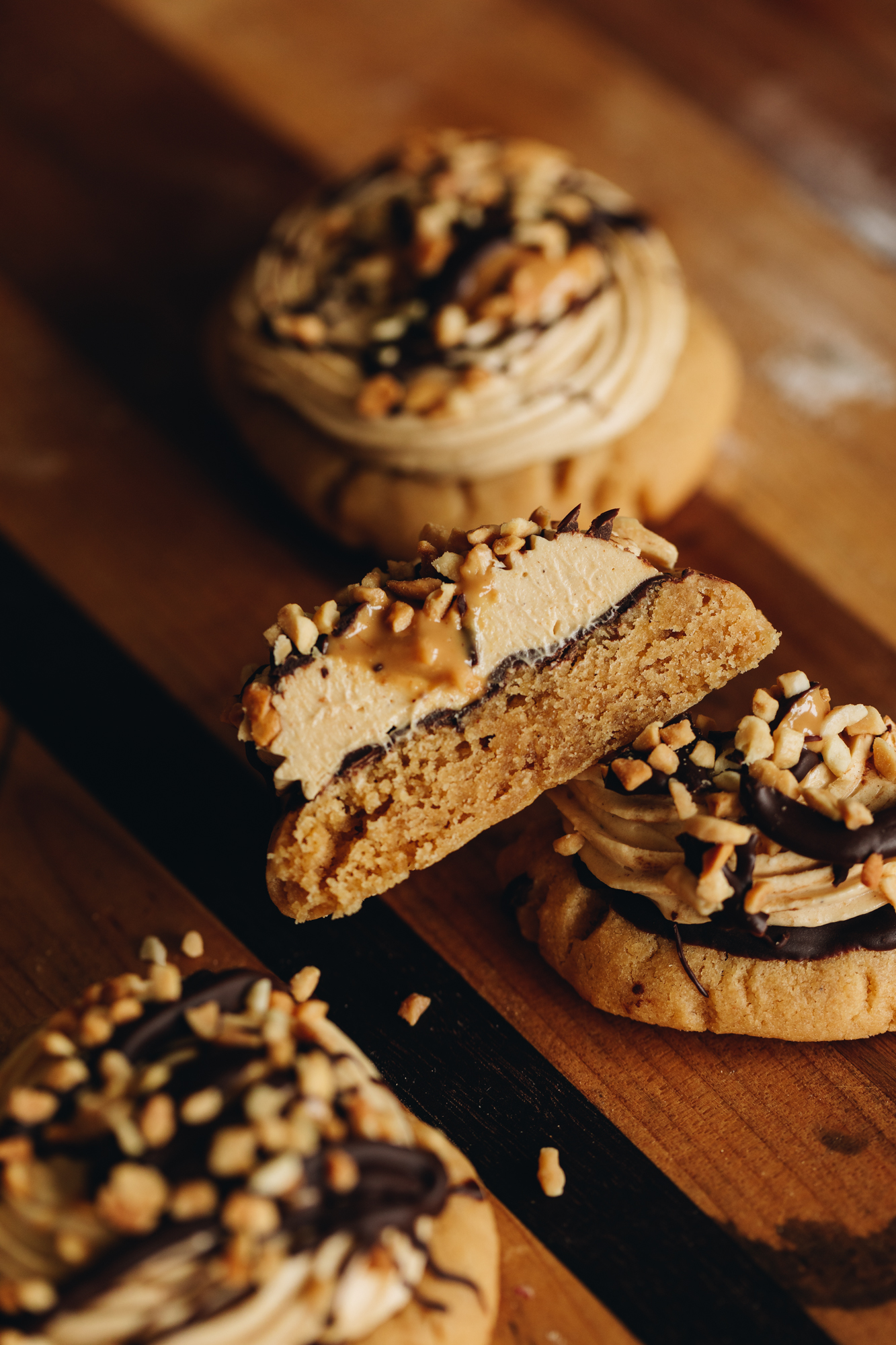 On a striped wooden board sits Ultimate peanut butter cookies that have been topped with piped peanut butter buttercream. They have a drizzle of chocolate added chopped but son top. One is cut in half to show the creamy filling.