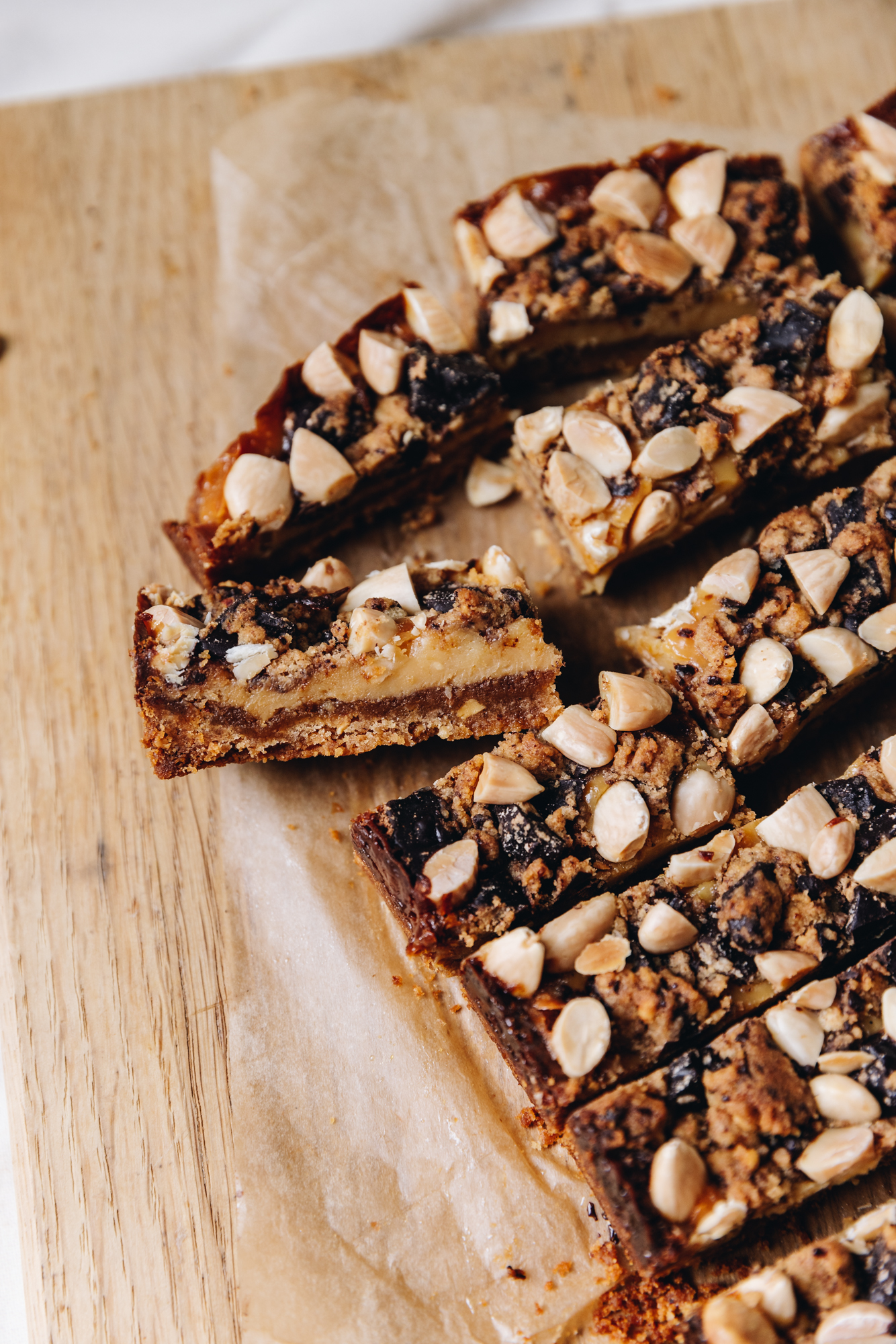 A wooden board is on a wooden table and is lined with brown baking paper. On it is slices of freshly baked caramel and almond slice. Once of the pieces has been turned over. The topping has chunks of dark chocolate and chopped almonds with biscuit crumb. The piece on its side shows the biscuit base, caramel filling and the topping. 