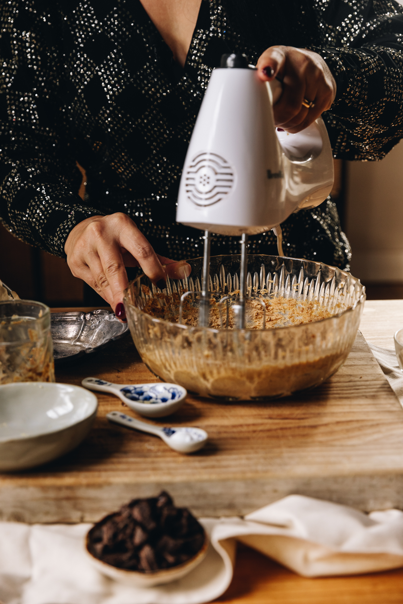 A wooden board sits on a cream cloth that is on a wooden table. On the board is a couple of empty vintage small bowls with nothing in it. It is also has two ceramic spoons on the board with baking powder in one. A glass vintage bowl is also on the board and it has whipped butter and sugar in it that is being whipped by a white hand mixer. 