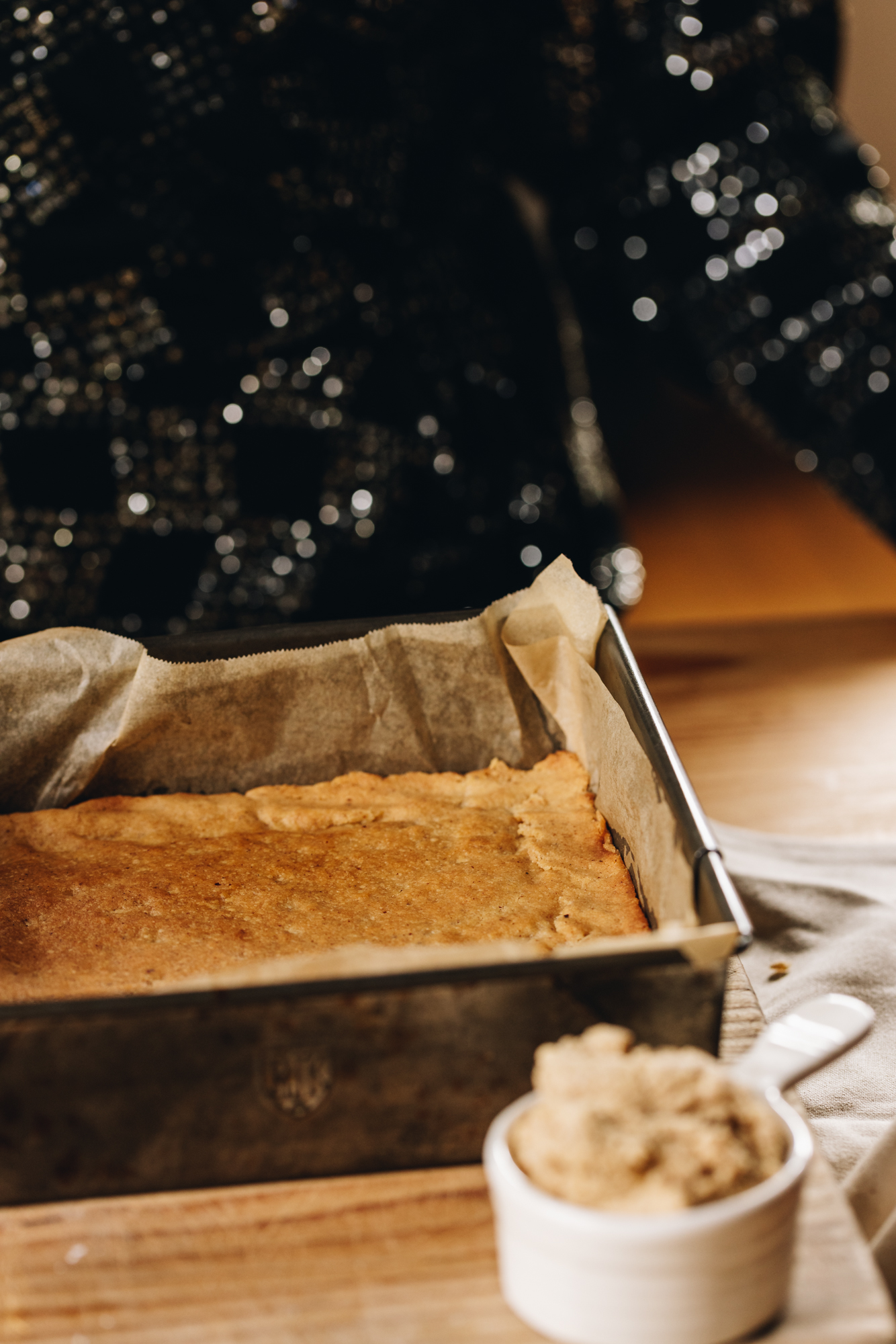 A vintage baking tray lined with brown baking paper is on a wooden board, sitting on a wooden table. In the tin is the baked base of the caramel and almond slice. There is a white ceramic cup of biscuit crumb in front of the tray too.
