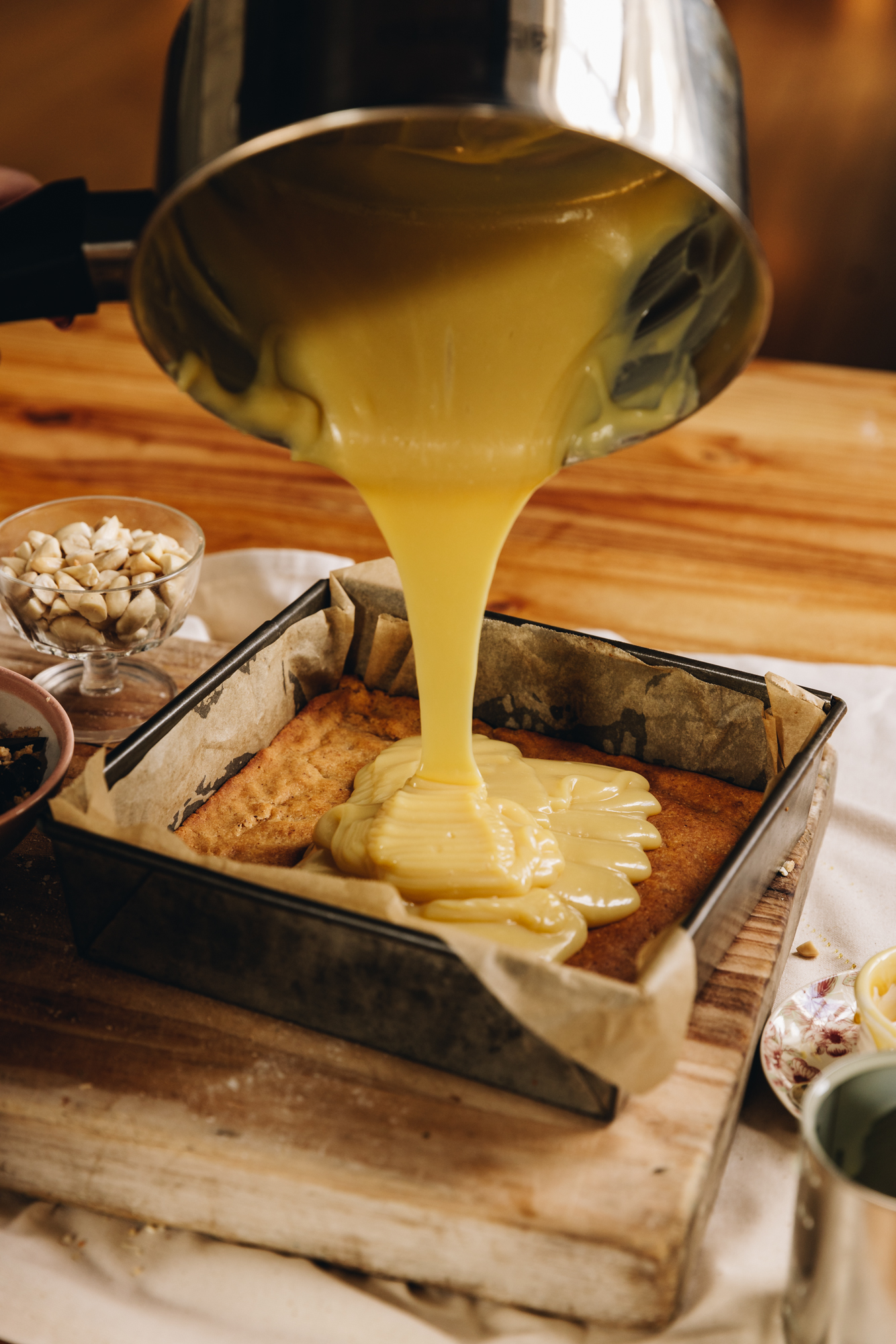 A vintage baking tray lined with brown baking paper is on a wooden board, sitting on a wooden table. The caramel and almond slice biscuit base is baked in the slice and a silver pot is pouring the caramel on top of it. Next to the baking tray is a glass vintage bowl is beached almonds and a small ceramic bowl with chocolate in it. 