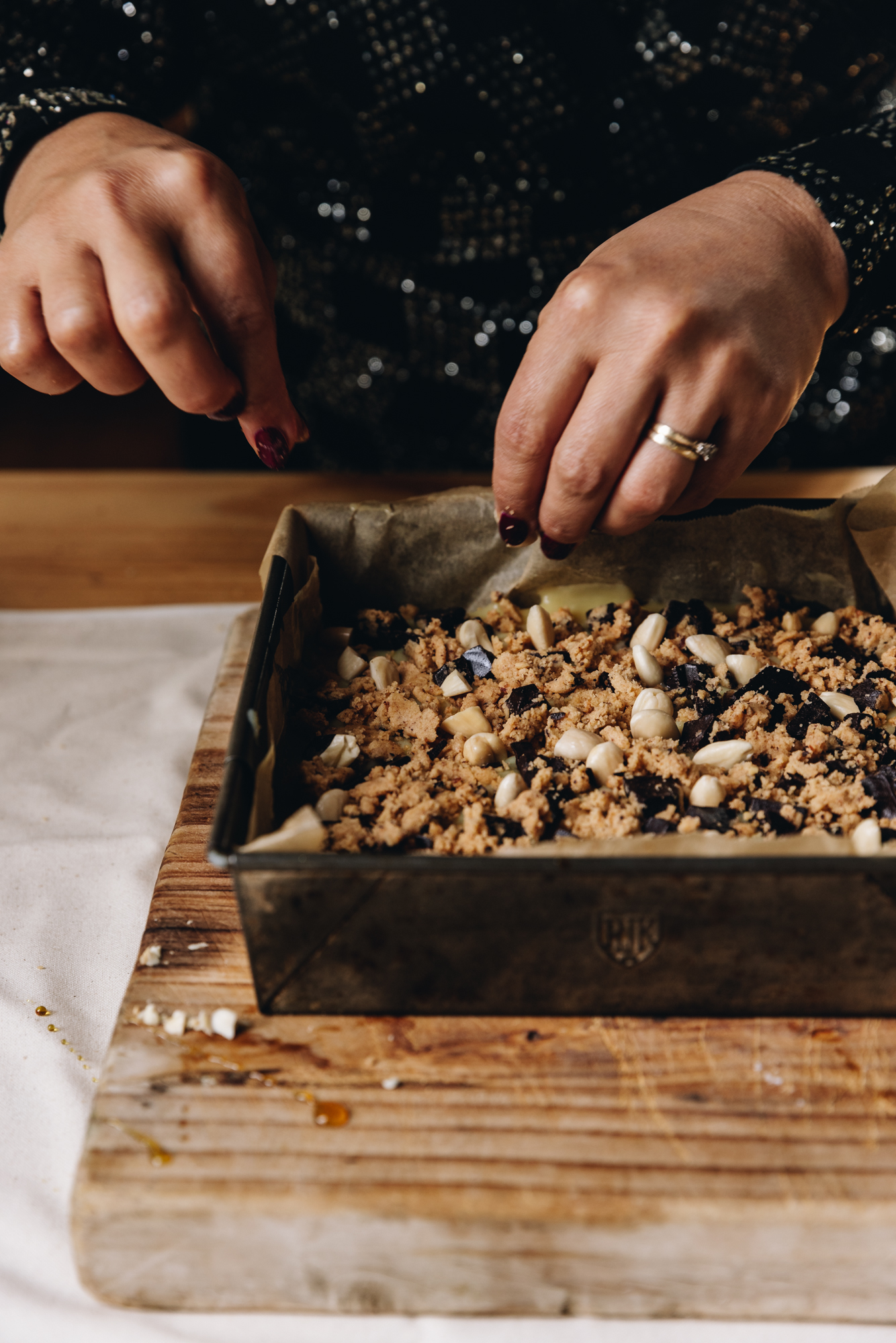 A baking tray sits on top of a wooden table that sits on a cream table cloth, on a wooden table. In the tin is a topping that has biscuit crumbs, dark chocolate chunks and blanched almonds. Naomi is adding some more almonds on top. 