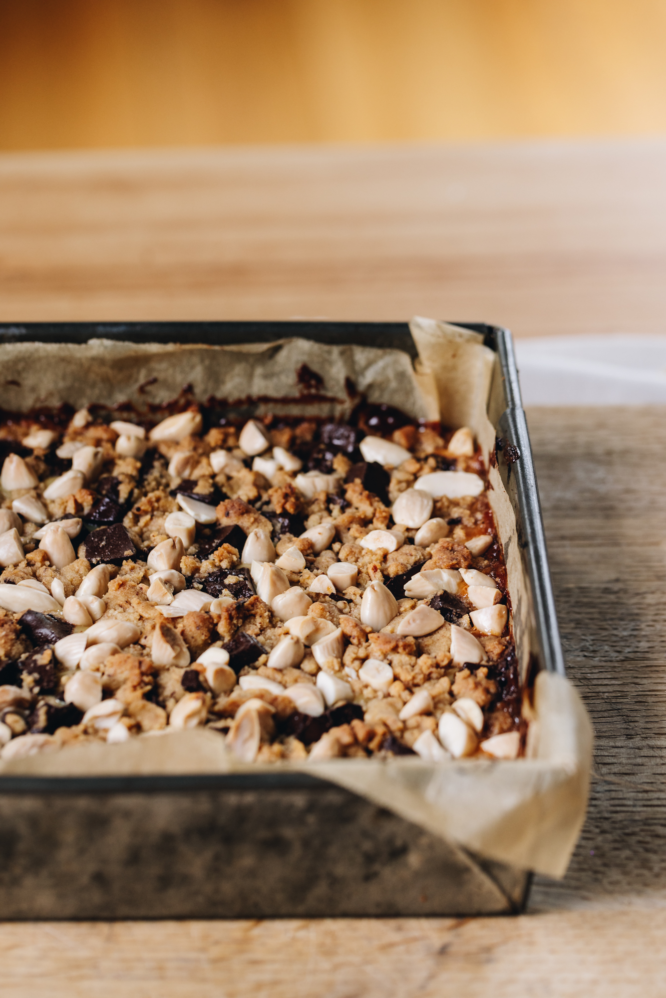 A baking tray sits on top of a wooden table that sits on a cream table cloth, on a wooden table. In the tin is a freshly baked caramel and almond slice that has biscuit crumbs, dark chocolate chunks and blanched almonds on top. 