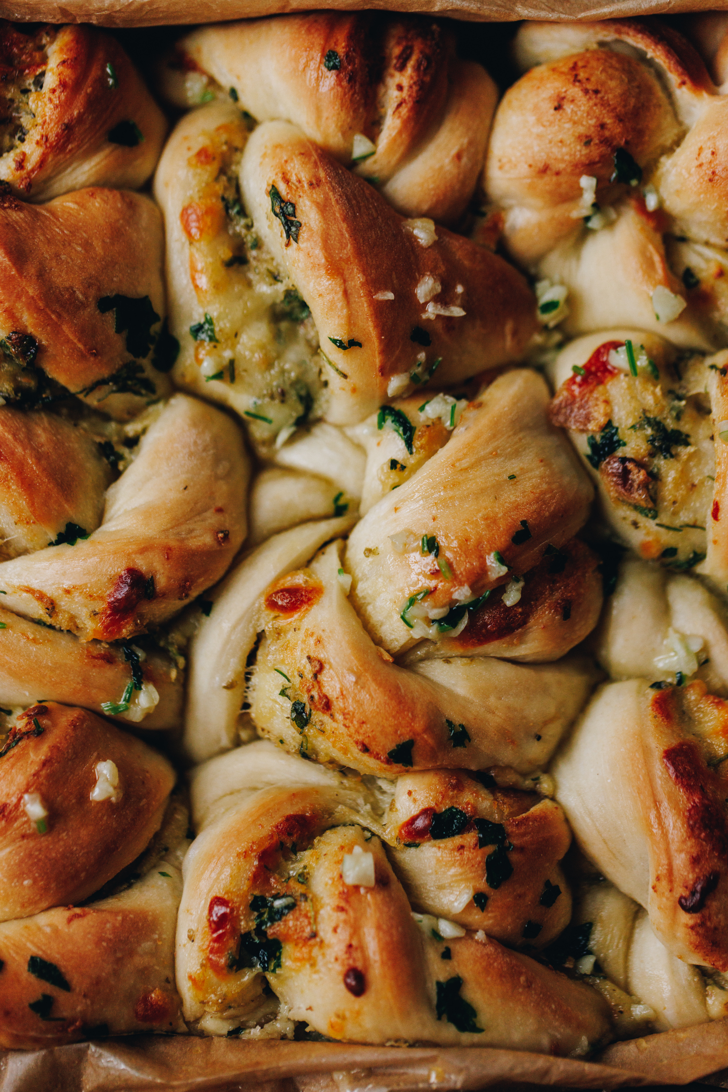 A close up, flat lay shot shows a photo of freshly baked roasted garlic knots with speckles of garlic and herbs seen in the dough and on top. 