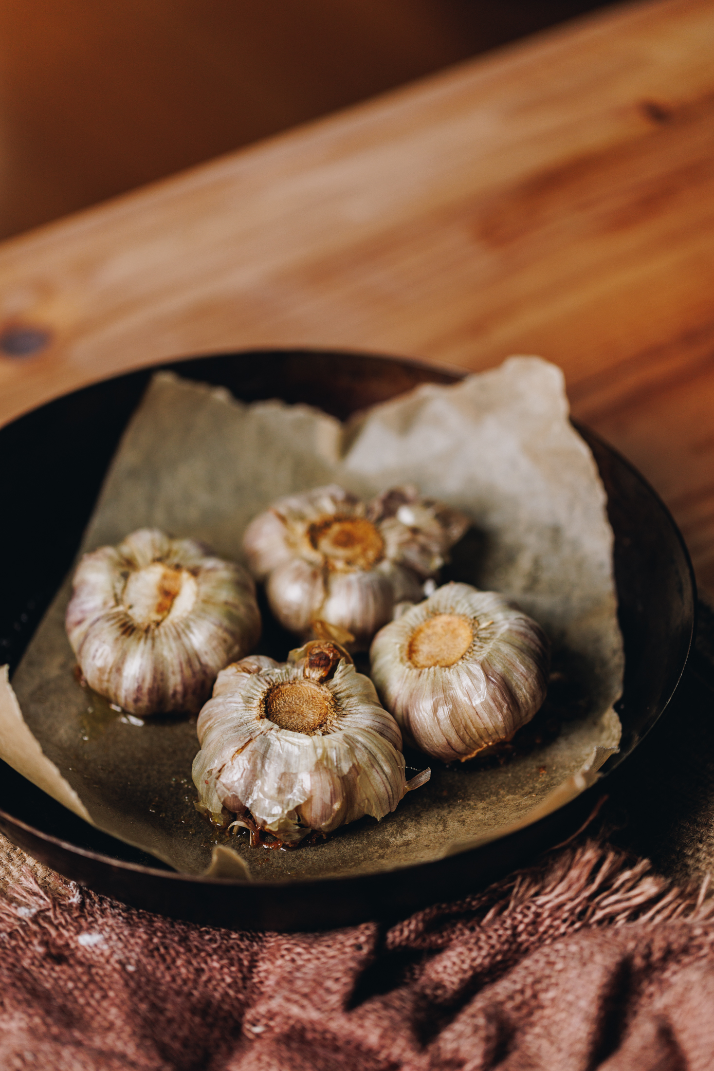 On a wooden table site a cast iron pan lined with baking paper. Four whole garlic cloves have been roasted are upside down on the baking paper. A pink natural tablecloth is seen at the front. 