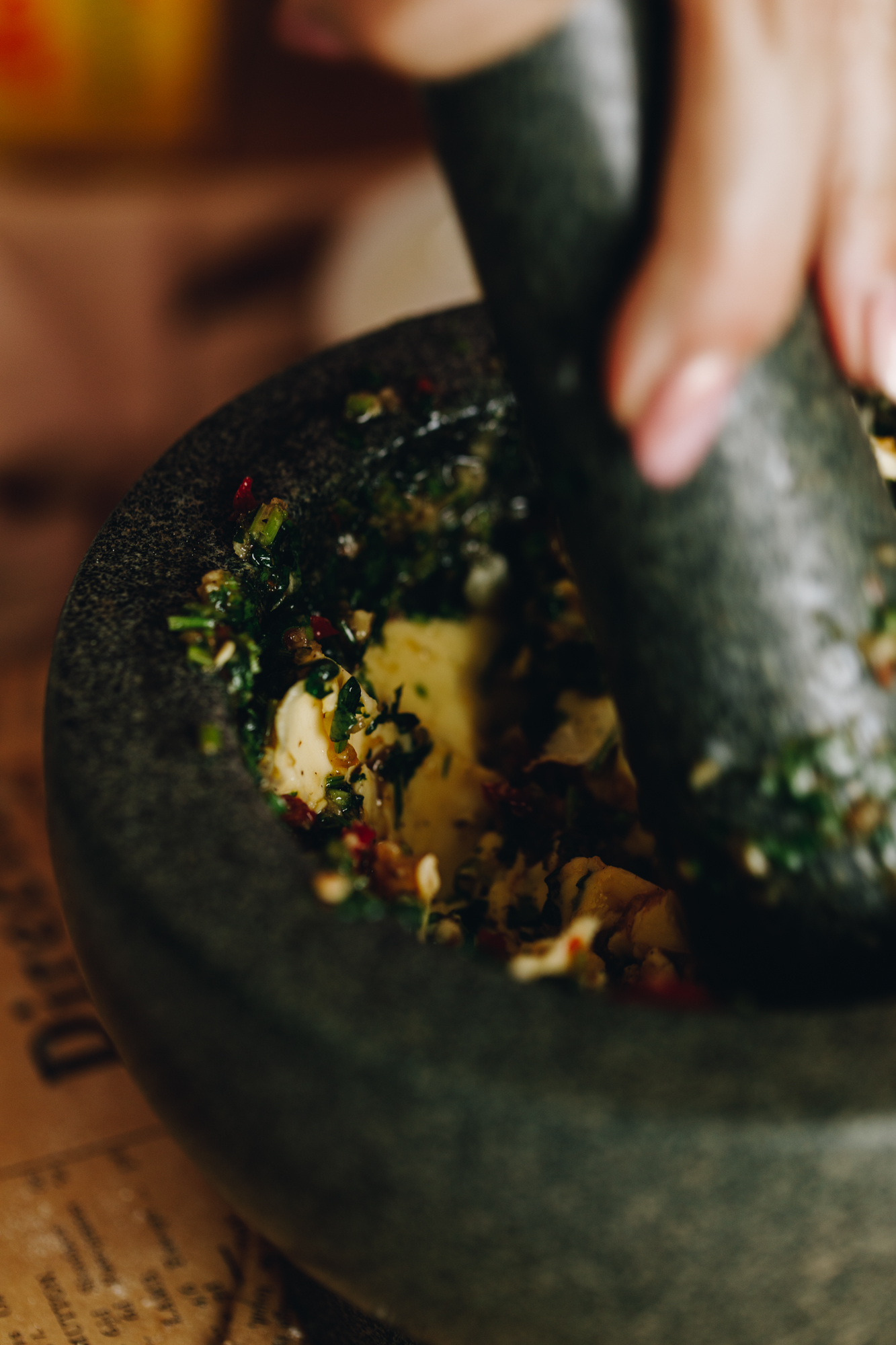 A charcoal coloured mortar and pestle is squashing a butter and herb mixture. The shot is close and speckled of fresh herbs are seen. 