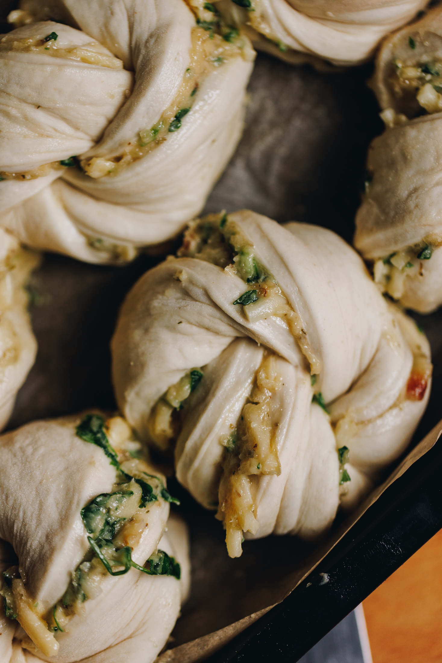 In a black tray lined with brown baking paper is unbaked roasted garlic knots that are filled with a garlic butter and herbs which are seen coming out of the unbaked dough. 