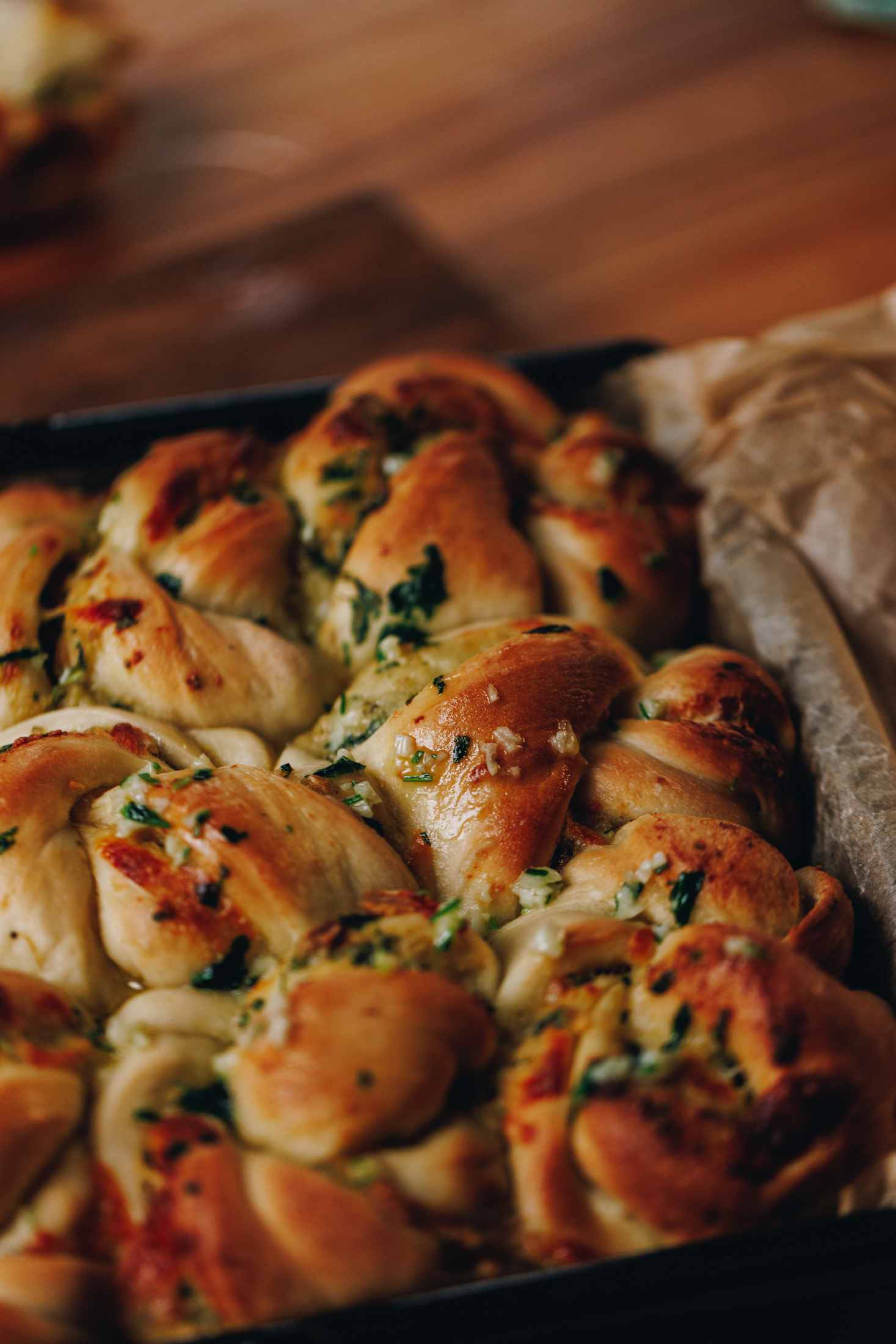 In a black tray lined with brown baking paper are freshly baked roasted garlic knots with speckles of garlic and herbs seen in the dough and on top. They are on a wooden table. 