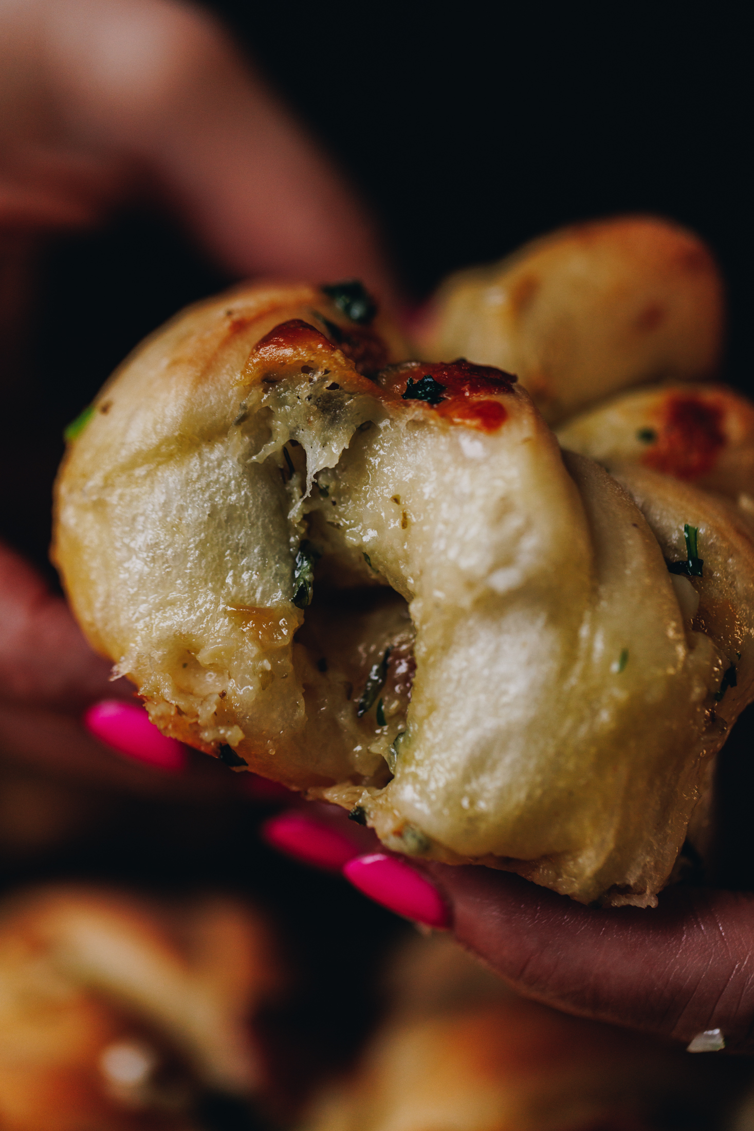 A extreme close up shows Naomi's hands holding a freshly baked roasted garlic knot showing the texture of the bun and the herby garlic butter peeking through. 