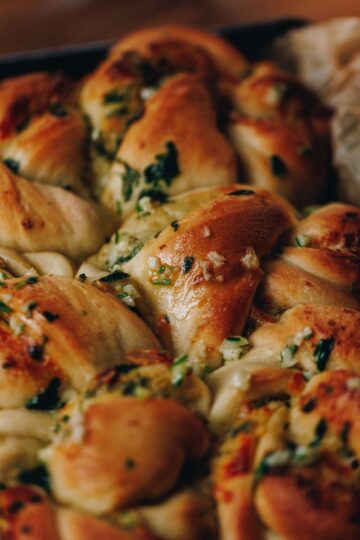 Freshly baked roasted garlic knots are in black tray with brown baking paper. They are shiny with butter and fresh herbs and filling are seen on the top of the knots.