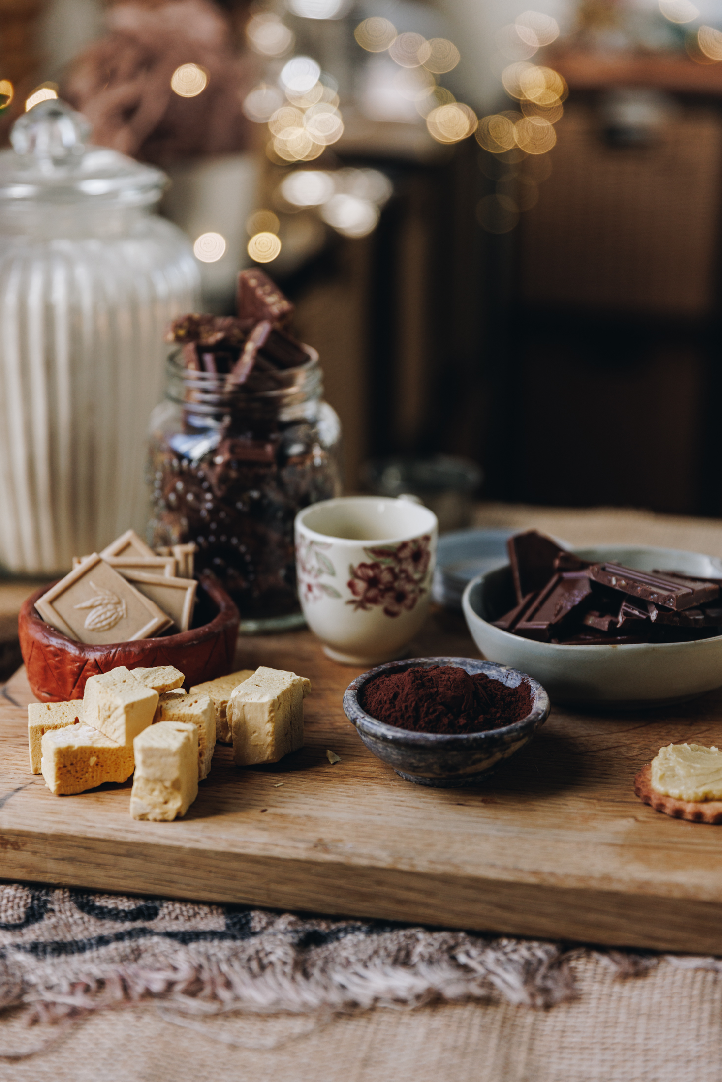 On a wooden table is natural fabric with a wooden board filled with biscuit ingredients.In view is honeycomb hunks, cocoa, dark chocolate, caramelised white chocolate and a jar of flour. Fairy lights are blurred in the distance.