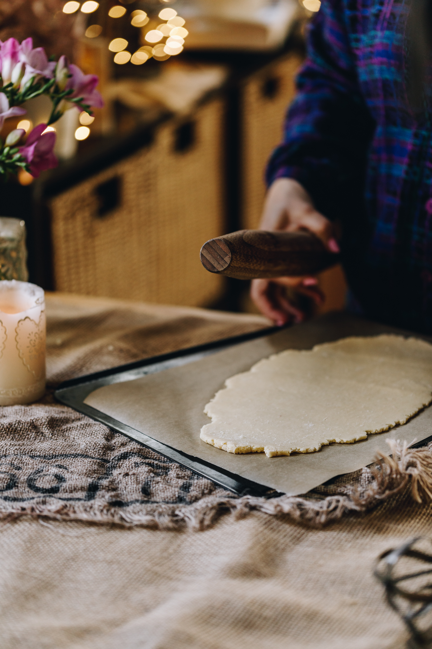 On a wooden table is natural fabrics. A black tray is on top of the fabric, lined with brown baking paper. On the paper is freshly rolled biscuit dough. Naomi is standing behind the tray holding a wooden chopping board. A candle burns next to her and fairy lights are blurred in the distance.