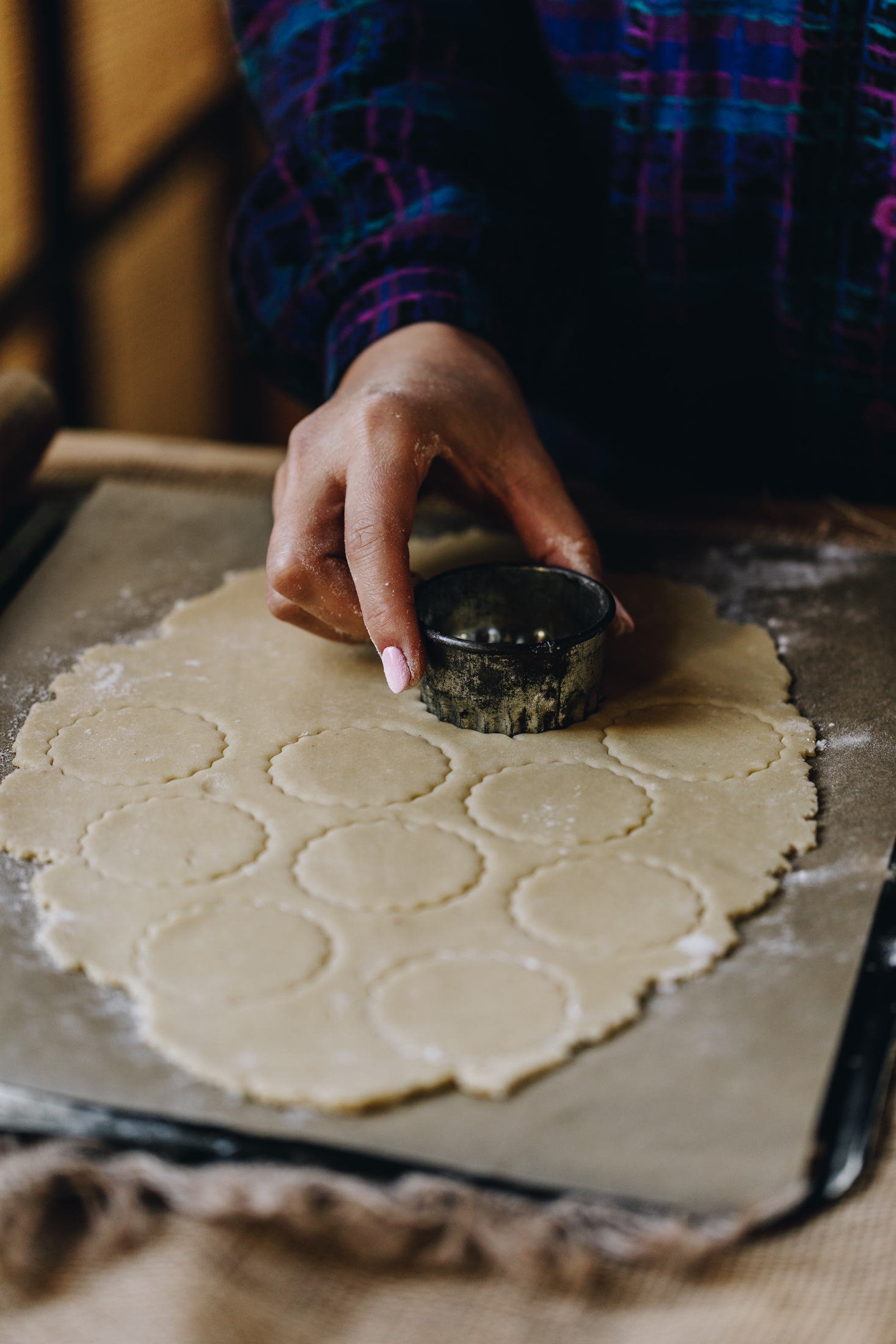 On a wooden table is natural fabrics. A black tray is on top of the fabric, lined with brown baking paper. On the paper is freshly rolled biscuit dough that has been cut out with a vintage cookie cutter that Naomi is holding as she presses it in to the dough.