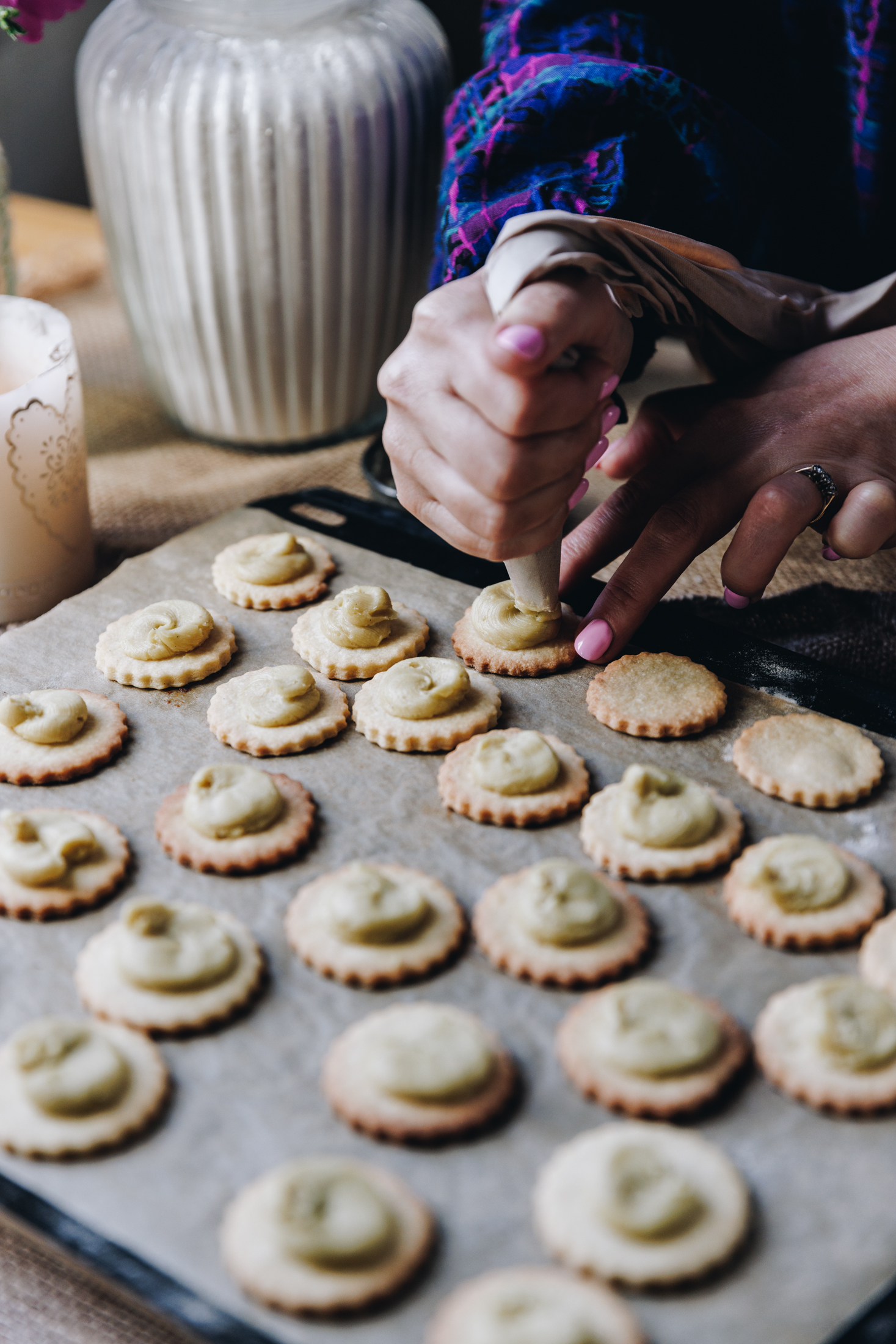 On a wooden table is natural fabrics. A black tray is on top of the fabric, lined with brown baking paper. On the paper is freshly baked shortbread biscuits that have buttercream piped on to them. Naomi is piping the second last biscuit with buttercream. A candle burns next to the tray and a glass jar of flour sits in the background.