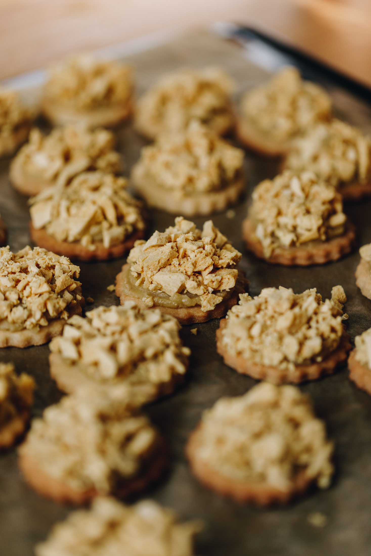 A black baking tray lined with brown baking paper sits on a wooden table. On the baking paper is shortbread cookies with buttercream on top and crushed honeycomb piled high on top of the buttercream.