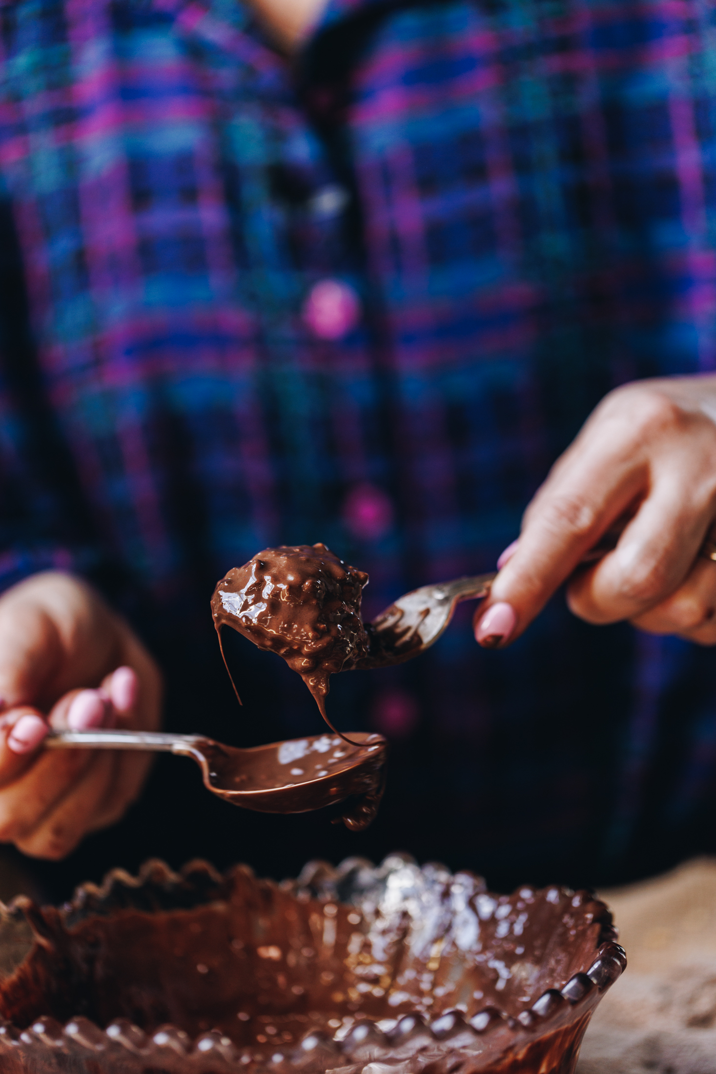 On a wooden table sits a purple vintage bowl filled with melted chocolate. Naomi is holding a fork with a homemade squiggle top biscuit on top of it. It has been drenched in chocolate. In the other hand Naomi is holding a spoon which is catching the drips of chocolate.