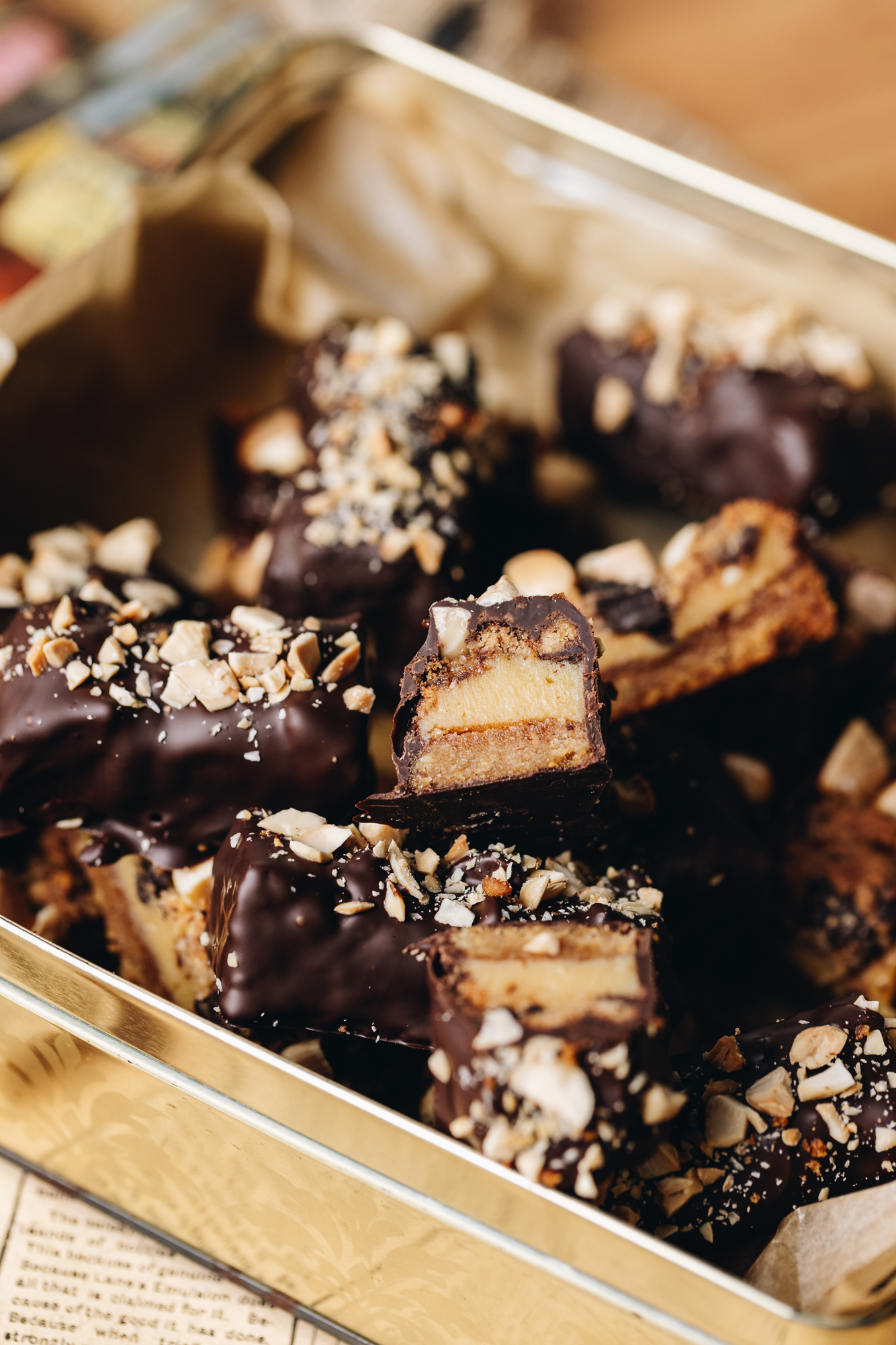 A golden baking tin sits on natural fabric on a wooden table. In the tin is caramel and almond chocolate bars. Some are covered in dark chocolate and have almonds sprinkle don top. Some of them have been cut open, showing the biscuit base, caramel filling and textured top.