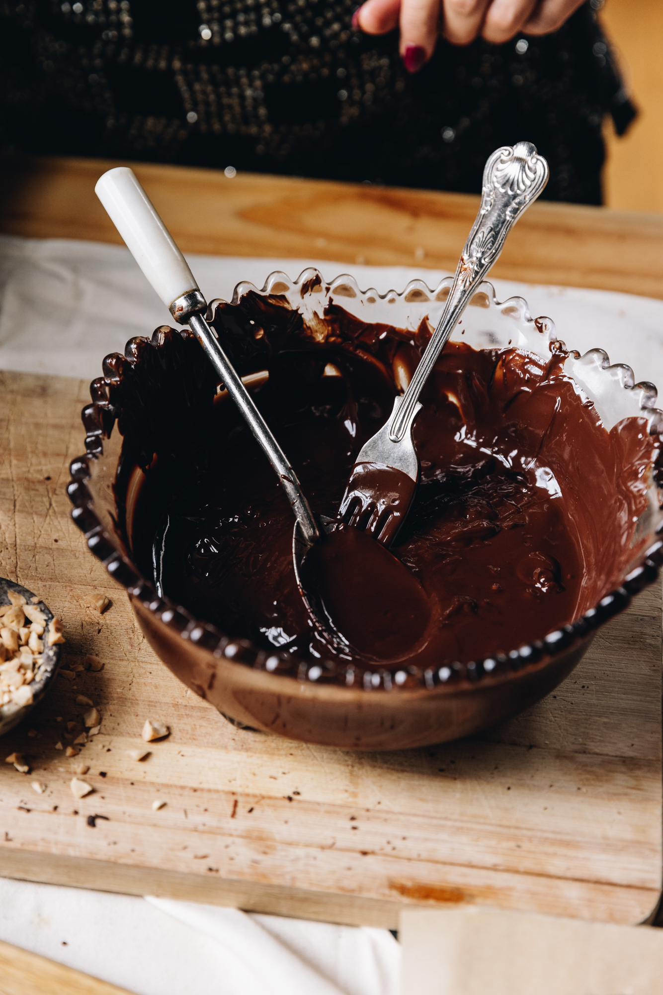 A wooden board sits on a cream table cloth, on a wooden table. On top is melted dark chocolate with a vintage fork and spoon in it. Almonds are sprinkled around it.