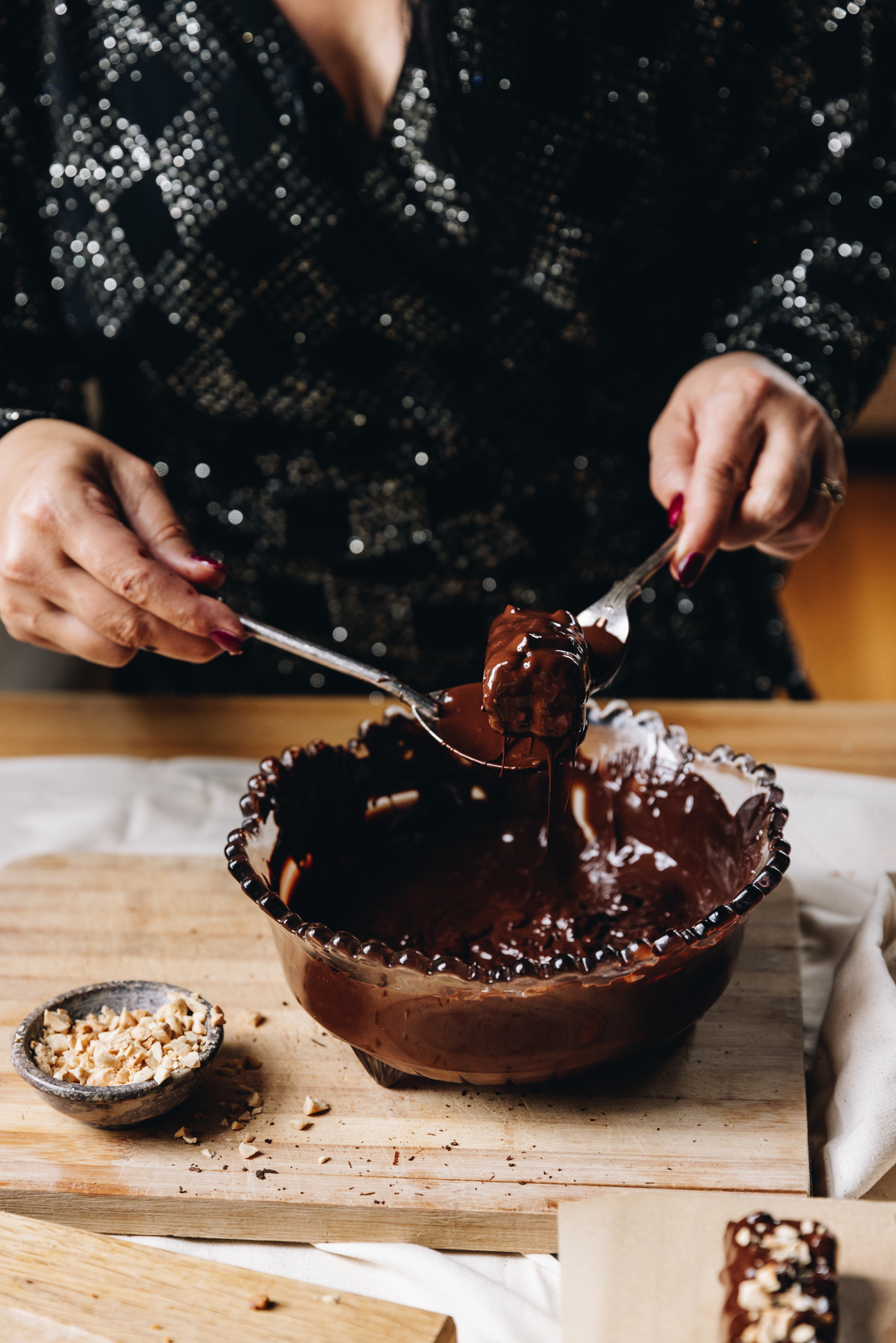 A wooden board sits on a cream table cloth, on a wooden table. On top is melted dark chocolate in a purple vintage bowl. Naomi is dipping a caramel and almond chocolate bar with a vintage fork and spoon. Almonds are in a small tin and sprinkled around it.