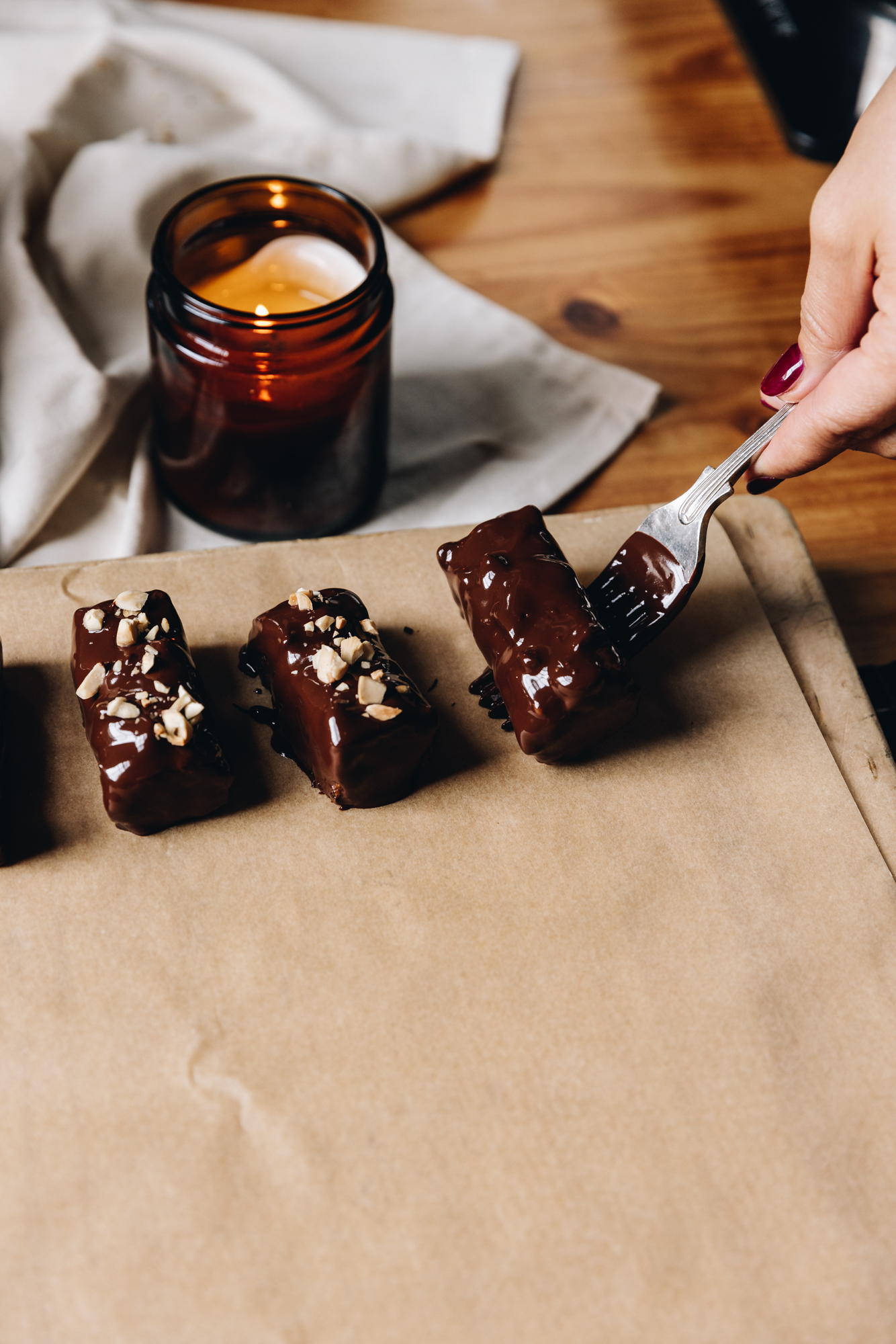 A wooden board sits on a wooden table with a cream tablecloth crunched behind it and a burning amber candle. On the board is brown baking paper, two pieces of freshly dipped caramel and almond chocolate bars are on the paper with crushed almonds on them. Another bar is being placed on the paper with a vintage fork, it has no decorations on top.