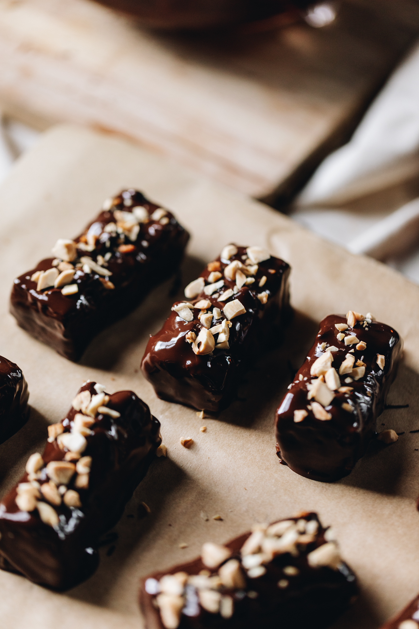 A wooden board is covered with brown baking paper. On the board is brown baking paper, six pieces of freshly chocolate dipped caramel and almond chocolate bars are on the paper with crushed almonds on them.
