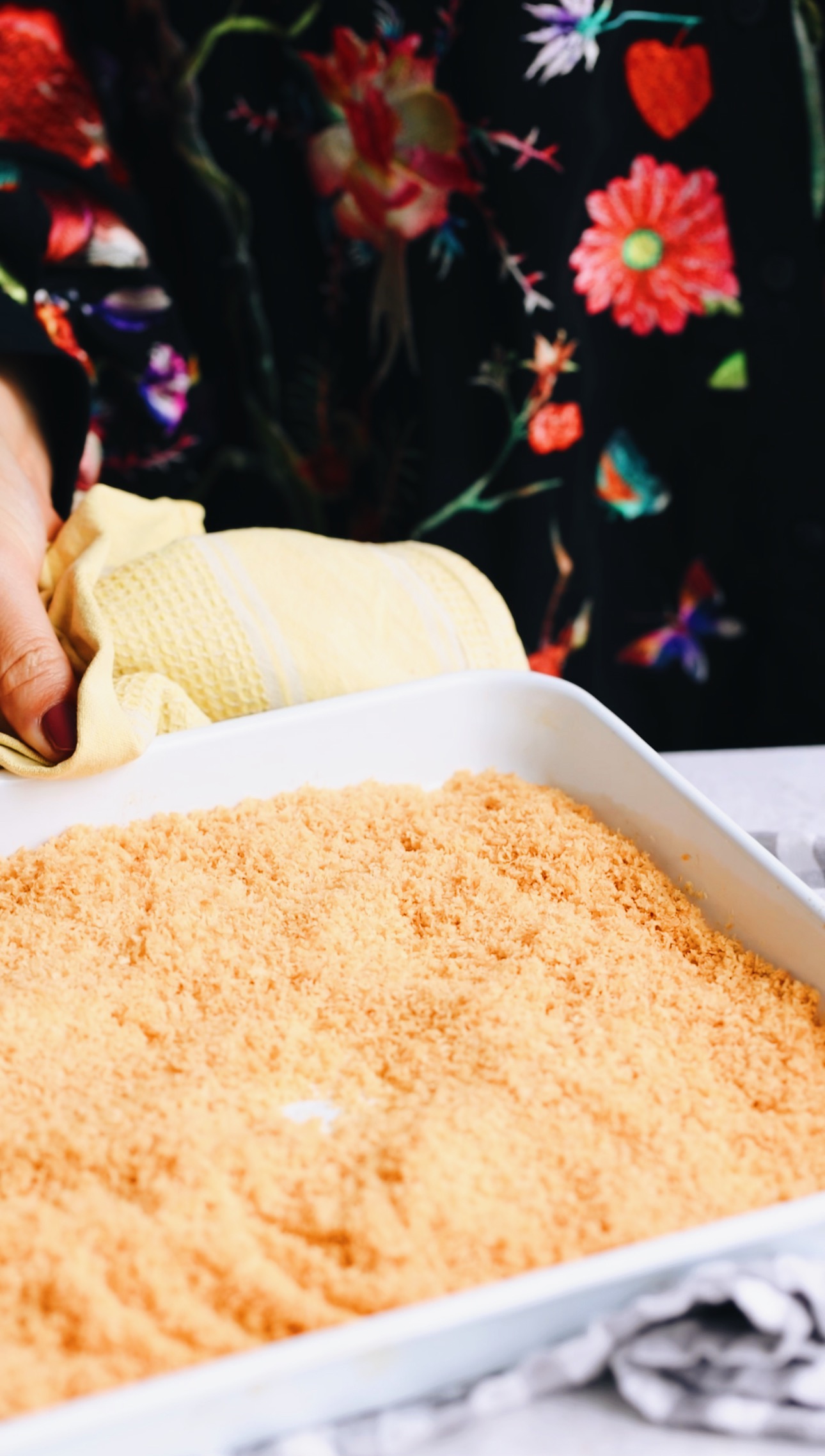 A white baking tray sits on top of a grey and white checked tea towel with golden toasted coconut in it. Naomi is placing it down and holding a yellow tea towel as she places it. 