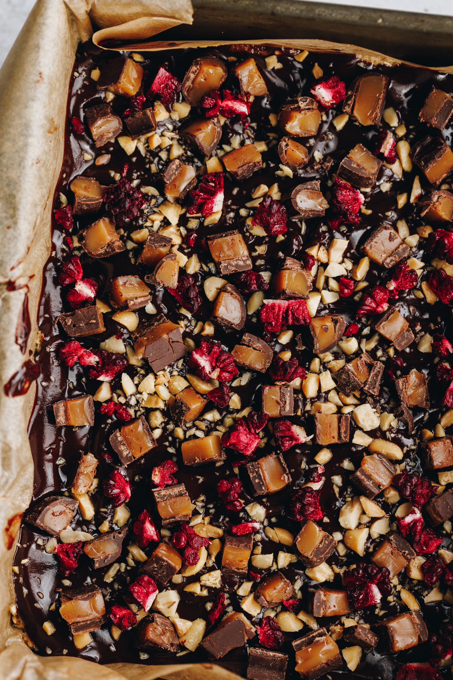A freshly decorated toasted coconut fudge sits in a baking tin lined with brown baking paper. The flat lay shows the texture of the topping. It has a chocolate topping with chopped chocolate covered caramels, chopped nuts and freeze-dried boysenberries on top. 