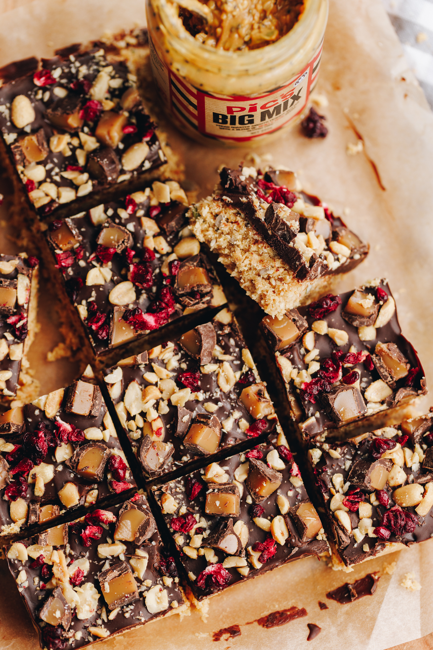 Toasted coconut fudge is decorated and sliced in to squares. It is on a board lined with brown baking paper. The flat lay shows the texture of the topping. It has a chocolate topping with chopped chocolate covered caramels, chopped nuts and freeze-dried boysenberries on top. One piece is flipped over to reveal the inside of the fudge. An opened jar of Pics Nut and seed peanut butter is on the corner too.