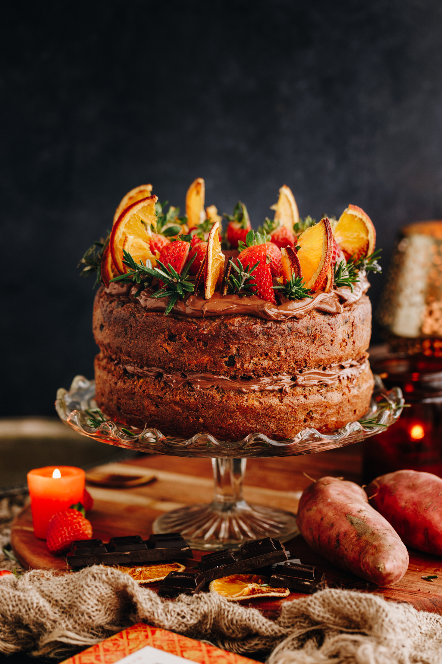 A glass cake stand sits on natural fabric on a table. On top of the cake stand is a kūmara and chocolate cake that has chocolate buttercream sandwiched in between the layers. It is has chocolate buttercream on top too and has slices of strawberries, dried oranges and rosemary on it. Under the cake stand is a small orange candle, a strawberry, orange slices, dark chocolate pieces and a couple of whole kūmara. An amber candle burns next to the cake. 