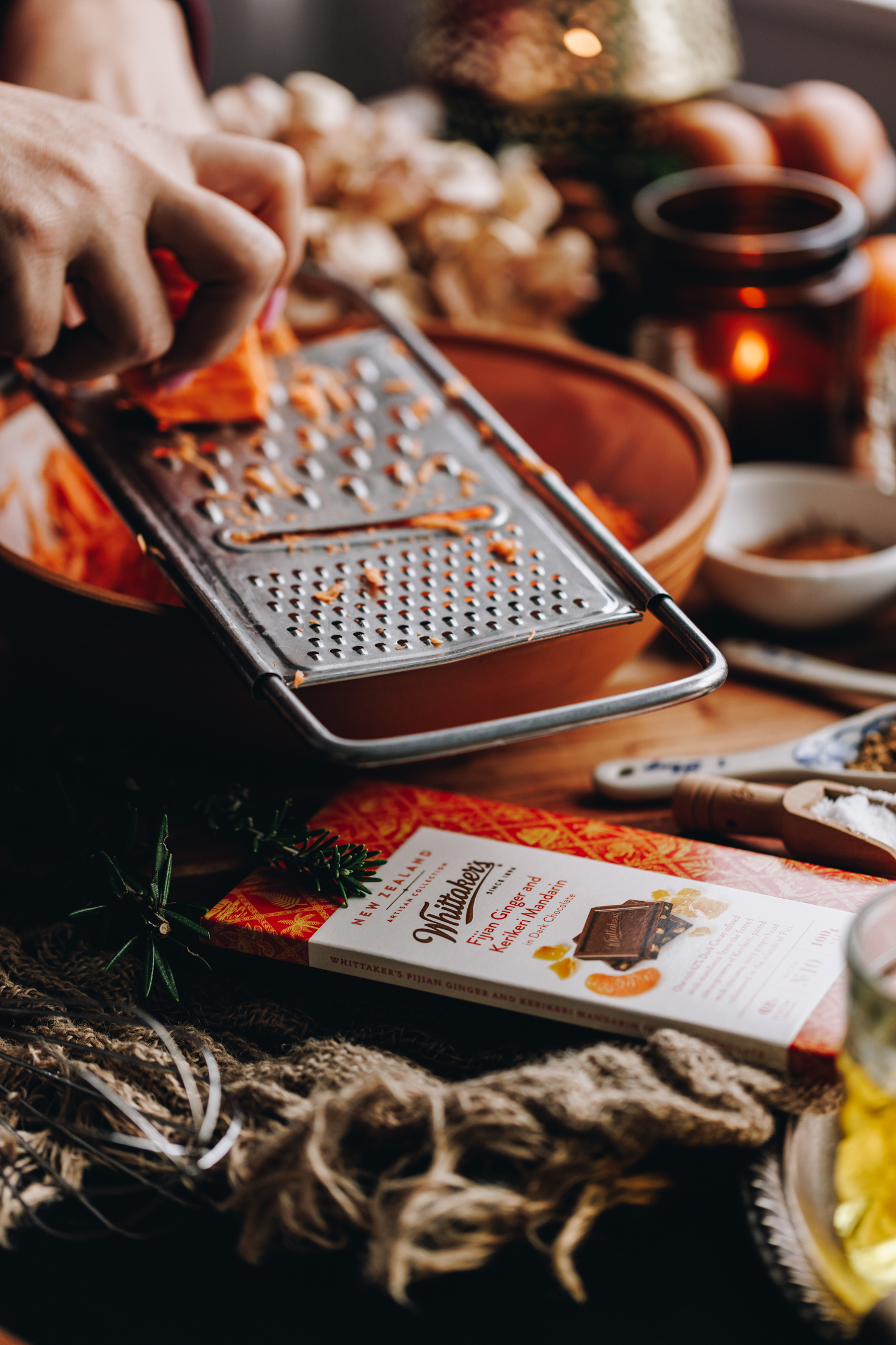 A natural piece of fabric sits on a table. A wooden board is on top of it and a block of Whittakers mandarin chocolate is nestled next to a ceramic bowl. There's a small wooden spoon with salt and a cermix teaspoon with spices. In the bowl is freshly grated kūmara and Naomi is grating more in to it. Behind the bowl is a small ramekin of spices, a burning amber candle and eggs. 
