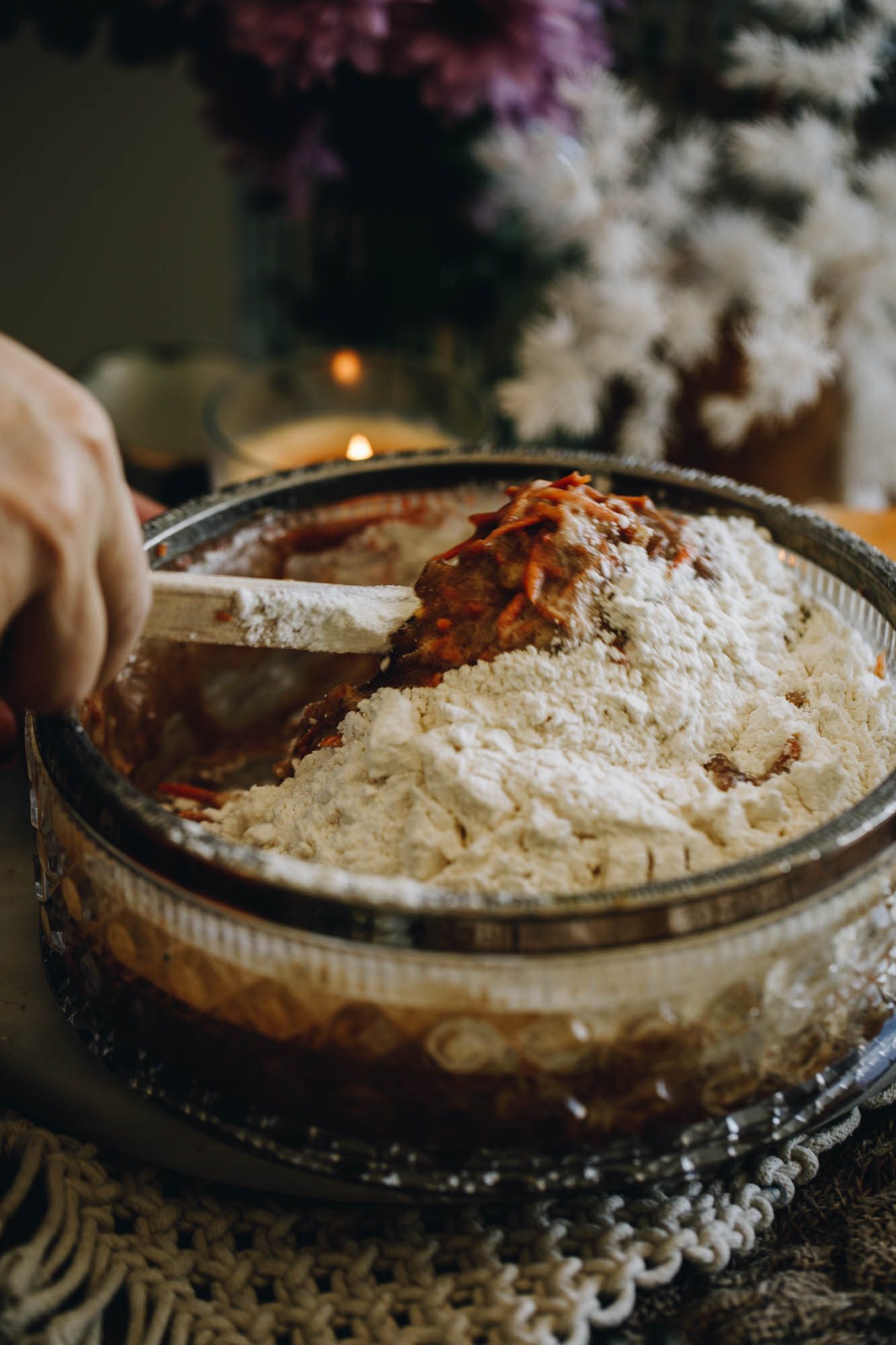 A glass vintage bowl with a silver trim sits on a weaved cream mat. In the bowl is kūmara cake mixture being stirring with a wooden spatula. Behind the bowl is a cream candle burning and white and purple flowers in a vase. 