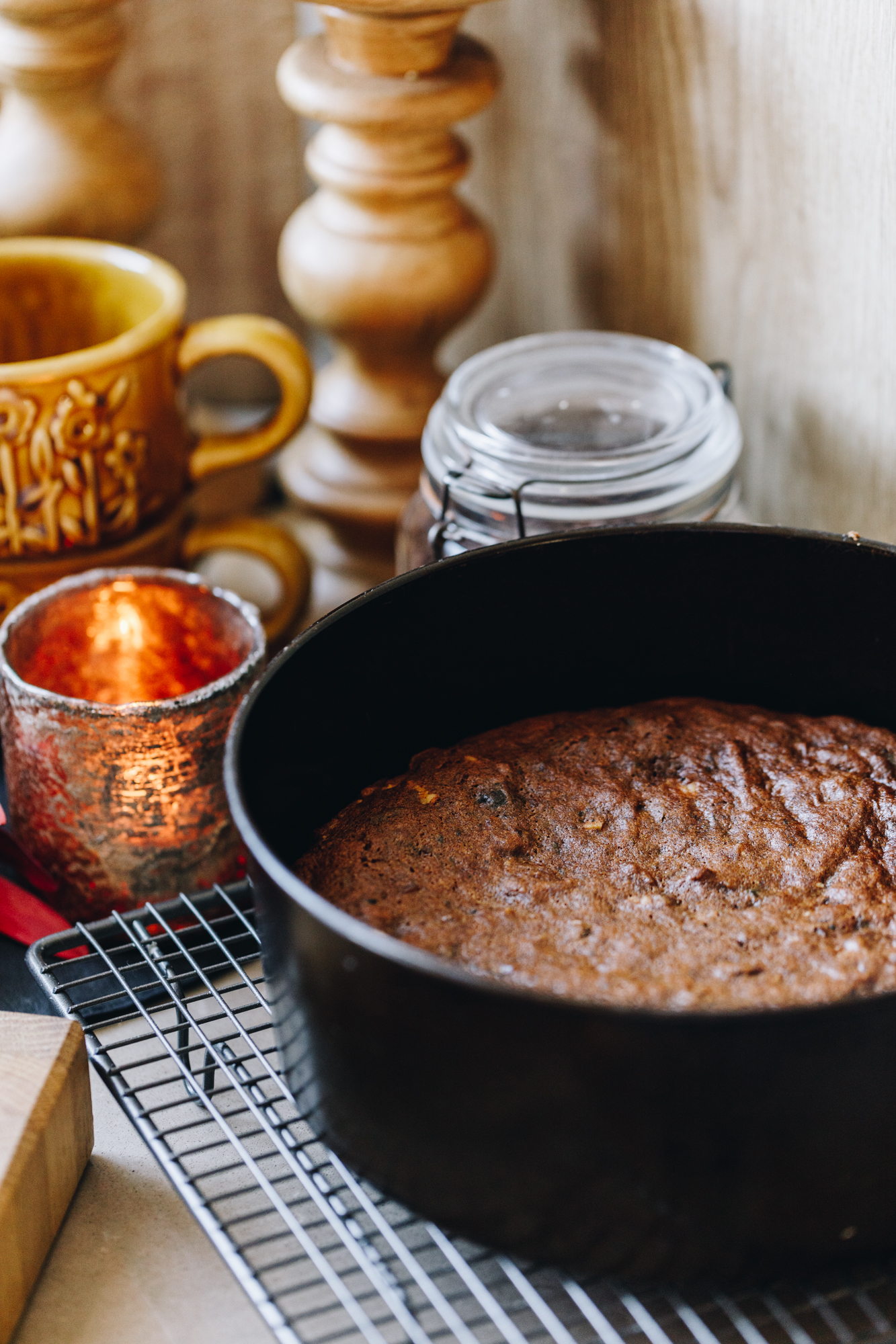A black cake tin sits on a wire cooling rack on a brown bench. In the cake tin is a freshly baked cake. Behind it is a burning orange candle, a jar, a pile of orange cups and wooden candlesticks. 