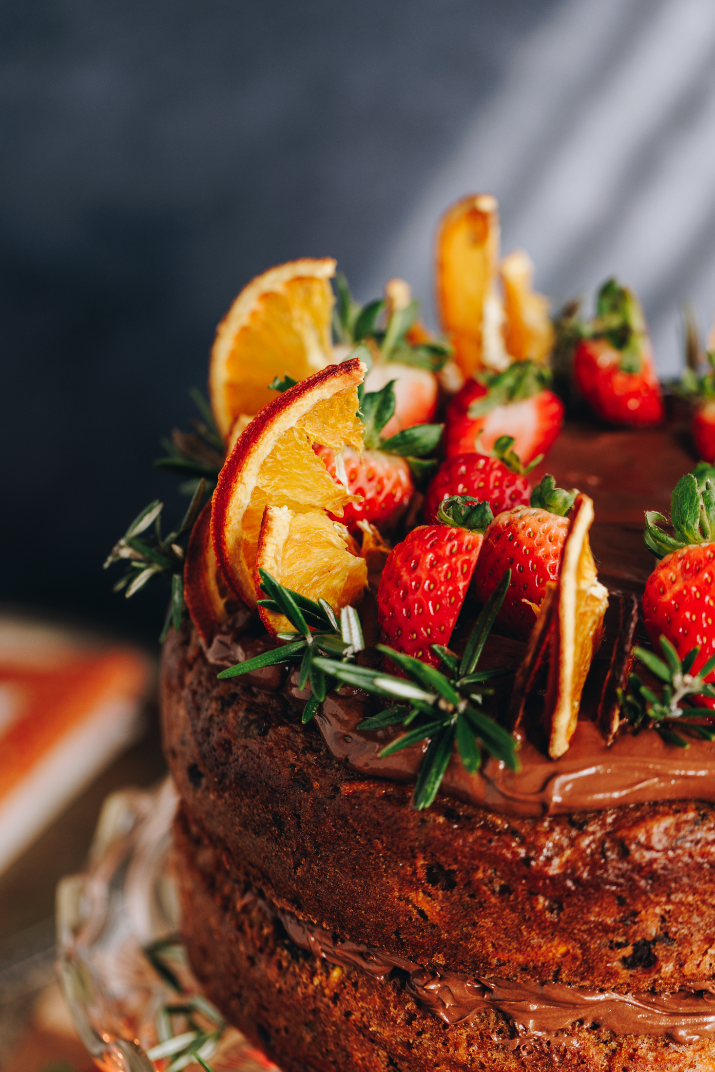 A close up shows a decorated kūmara and chocolate cake. It is has chocolate buttercream on top too and has slices of strawberries, dried oranges and rosemary on it. 