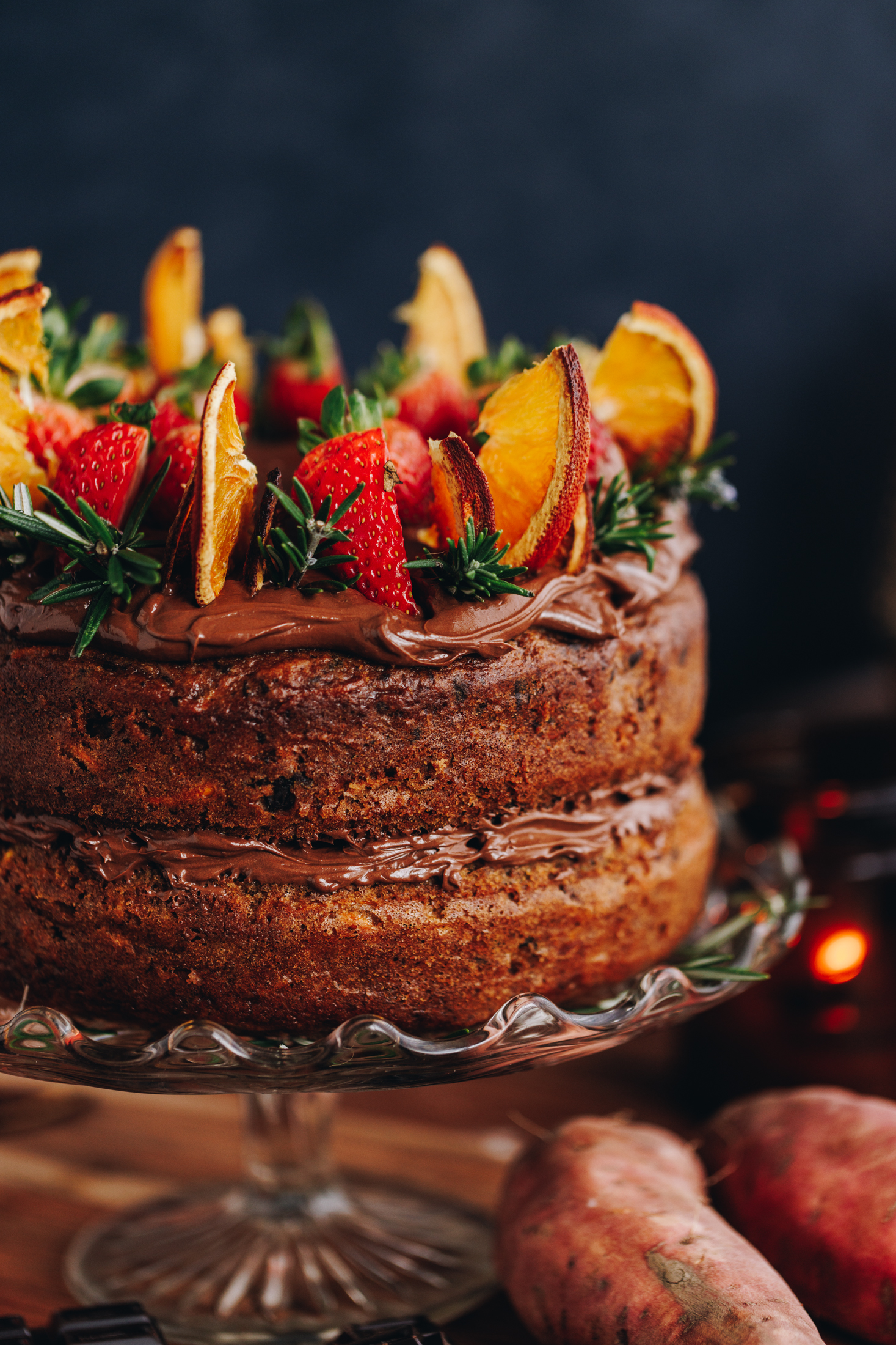 A glass cake stand sits on a wooden table. On top of the cake stand is a kūmara and chocolate cake that has chocolate buttercream sandwiched in between the layers. It is has chocolate buttercream on top too and has slices of strawberries, dried oranges and rosemary on it. Under the cake stand are a couple of whole kūmara.