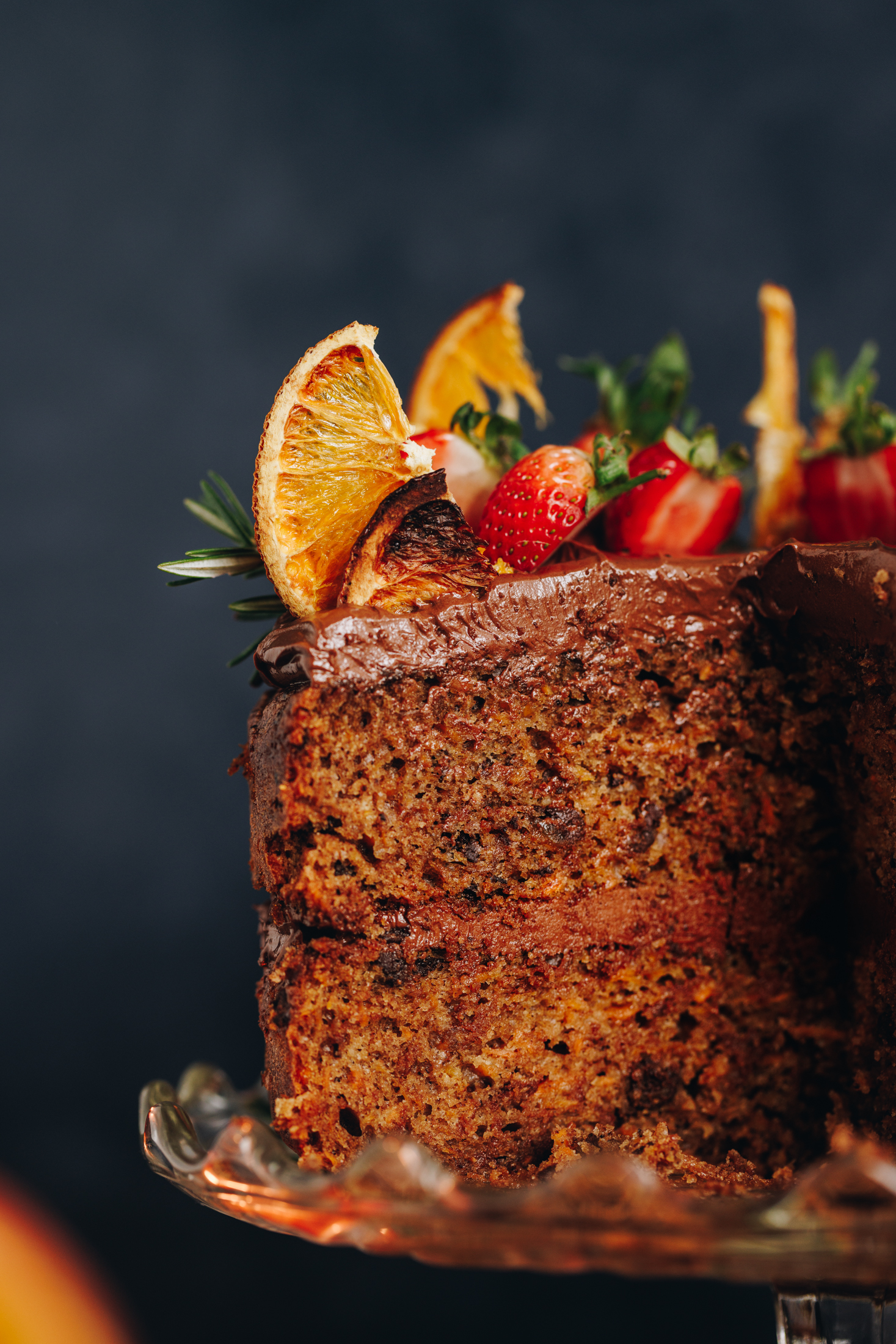A kūmara and dark chocolate cake is on a glass cake stand. It has been cut open to reveal the chocolate buttercream in between the cake layers. It is has chocolate buttercream on top too and has slices of strawberries, dried oranges and rosemary on it.