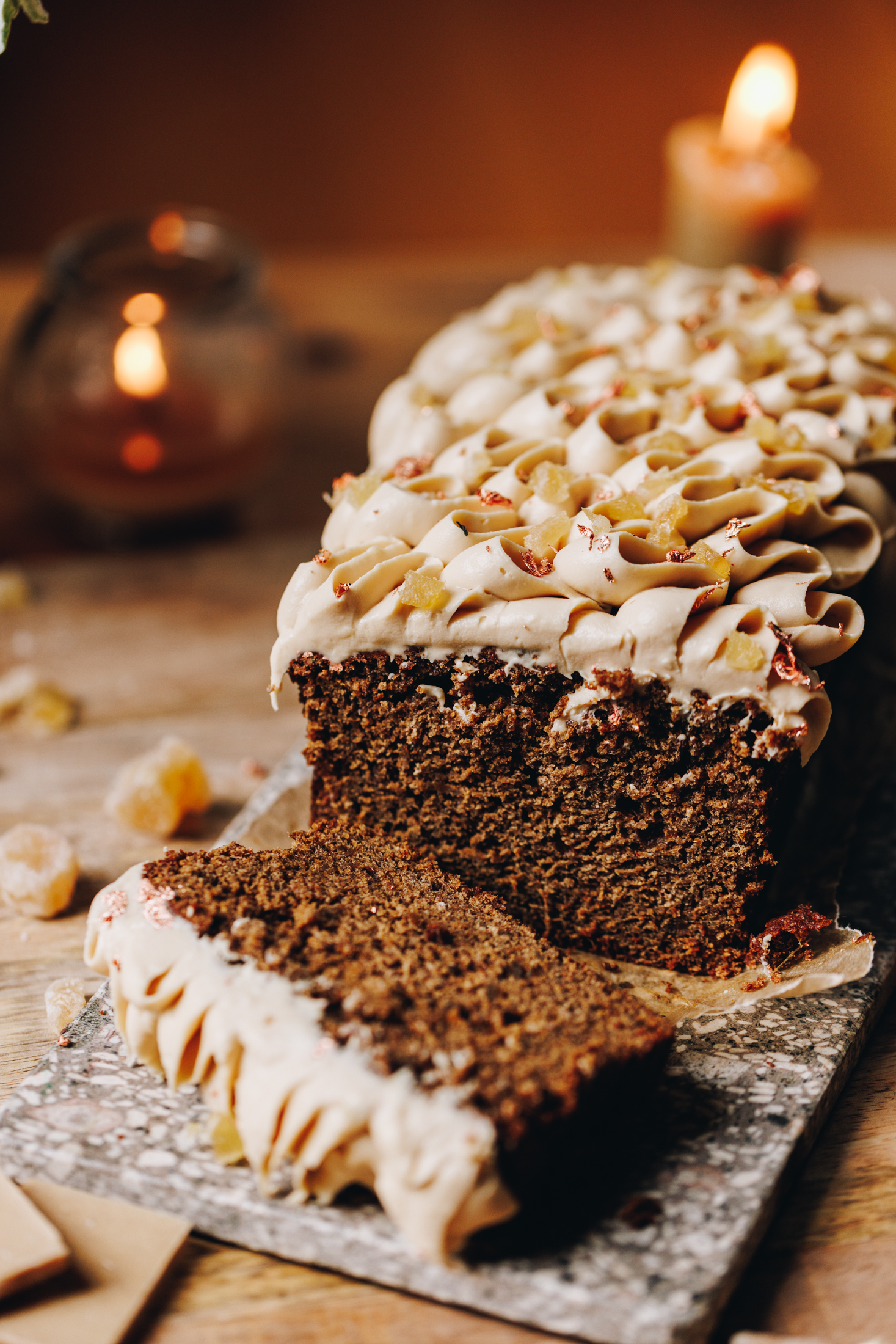 On a wooden table sits a small stone board. Two candles are burning in the background. On the stone board is a baked spiced gingerbread loaf that has piped white chocolate cream cheese frosting on it. A lice has been cut and is laying flat, showing the inside of the loaf.