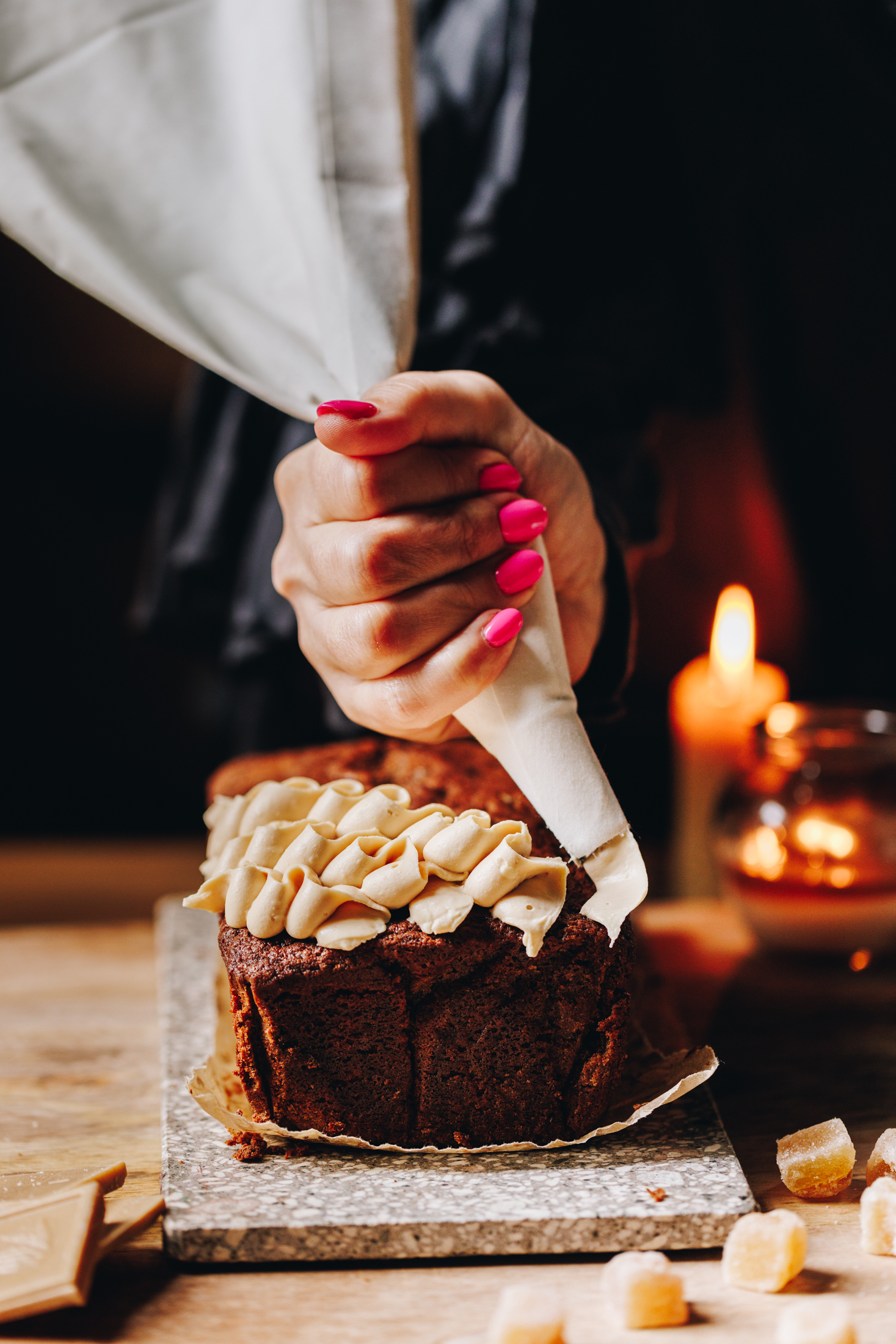 On a wooden table sits a small stone board. Two candles are burning in the background. On the stone board is a baked spiced gingerbread loaf that is having white chocolate cream cheese frosting piped on it in a squiggly pattern.