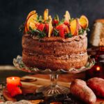 A kumara and chocolate cake is on a glass cake stand, it has been topped with dark chocolate buttercream, dried oranges and fresh strawberries. Orange kumara are next to it.