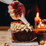 White chocolate cream cheese frosting is being piped on to a Ginger loaf in a squiggly pattern. Candles are in the background.