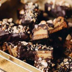 Caramel and almond chocolate bars are piled on top each of other, one is sliced open to reveal the inside of the bar showing the biscuit base, caramel inside and chocolate coating.