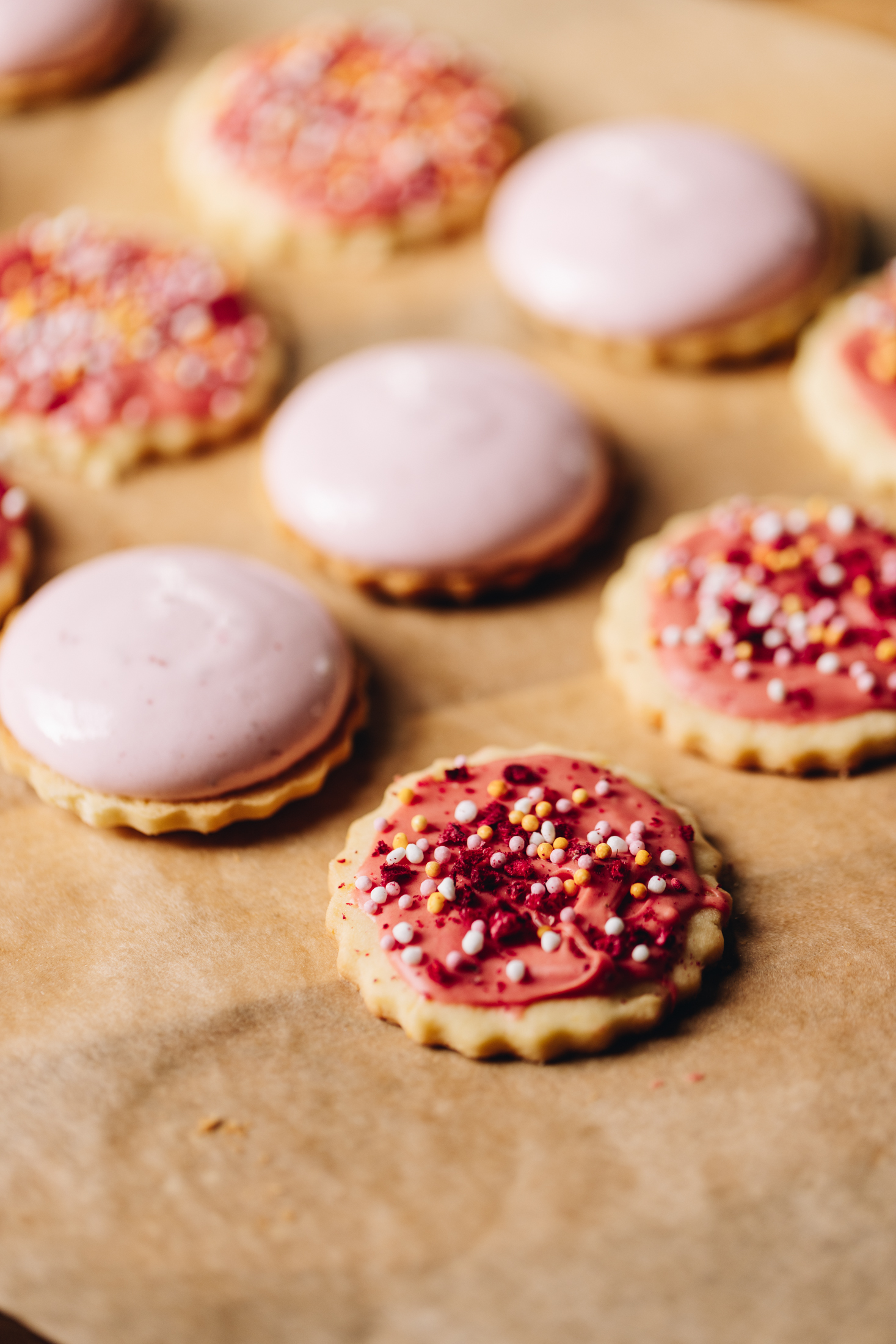On brown baking paper is hundreds and thousands biscuits baked and decorated, every second biscuit has pink raspberry marshmallow piped on to it. 