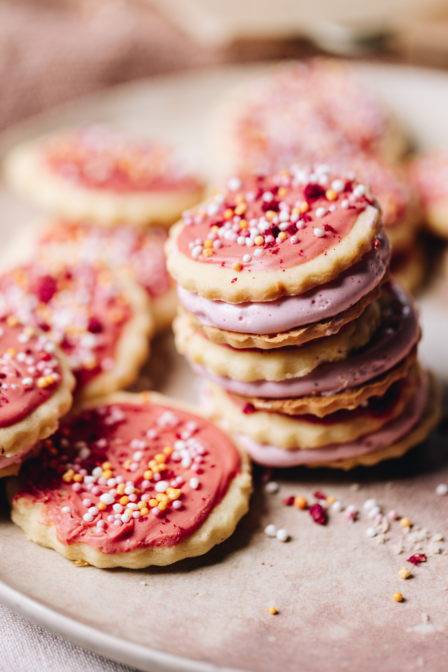 Hundreds and thousands biscuits are filled with marshmallow. There is three in a pile and others scattered around it on a pink plate. 