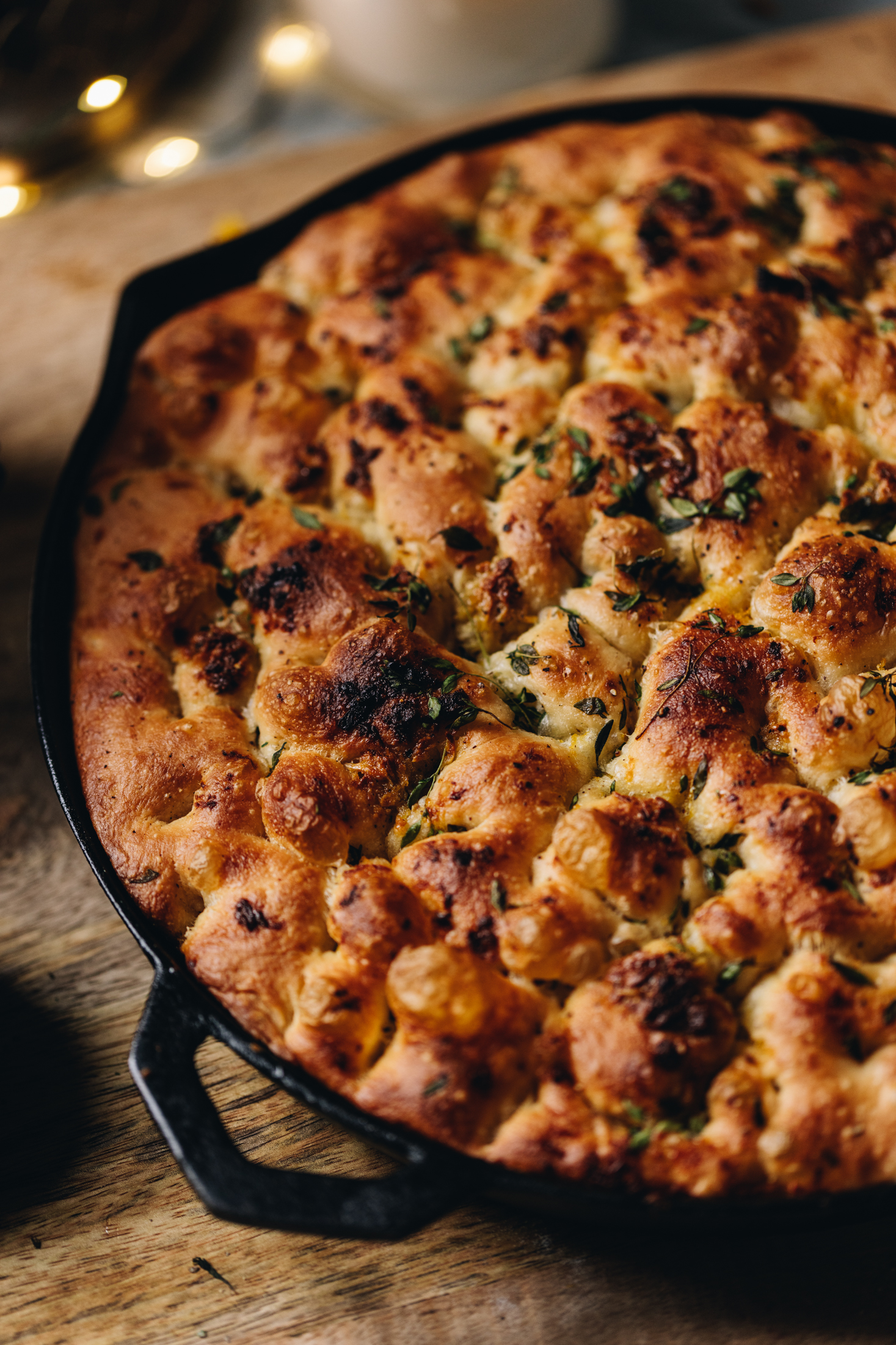 In a cast iron pan is a freshly baked no-knead focaccia bread. It is on a wooden table.