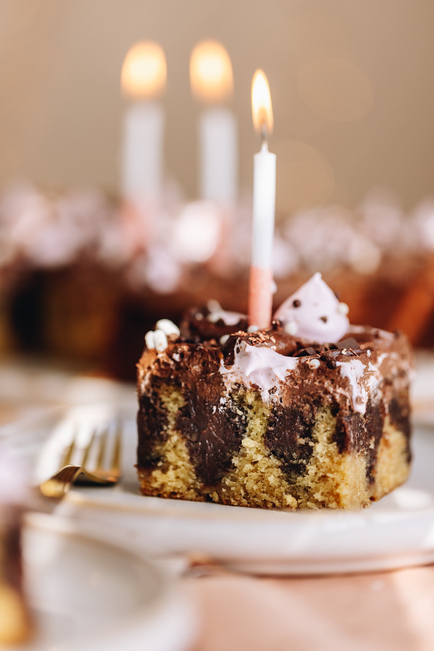 A white plate sits on a pink table cloth. On the plate is a slice of chocolate peanut butter poke cake. The slice shows the vanilla cake with chocolate ganache poked in to it. It is topped with a chocolate buttercream and pink blobs of meringue. A candle is burning in the centre of it. Behind the slice in the remaining cake blurred in the background with two candle burning behind it.