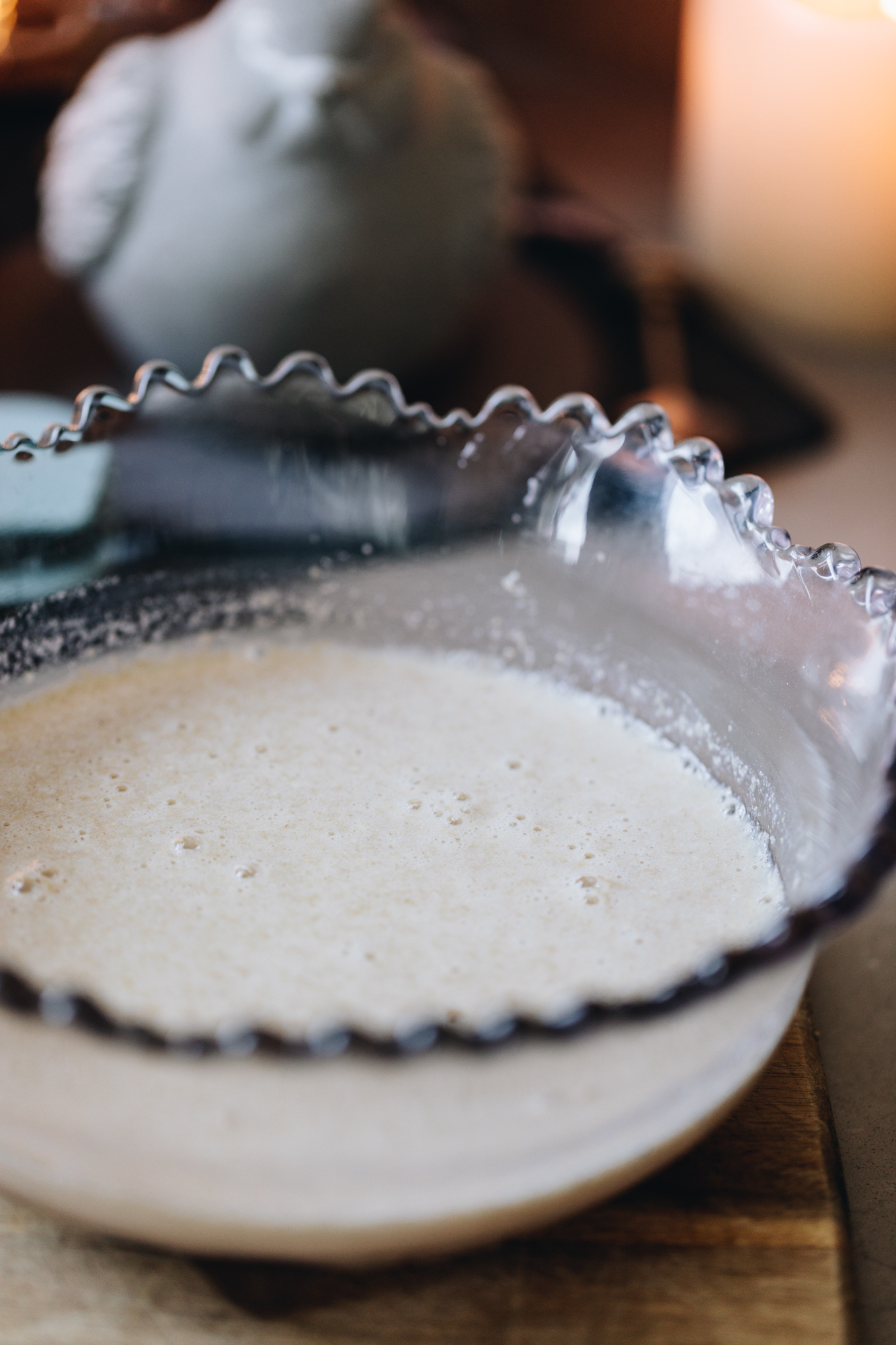 A light purple vintage bowl with a crinkled eade sits on a wooden board. In the bowl is homemade buttermilk. A ceramic bird is in the background.