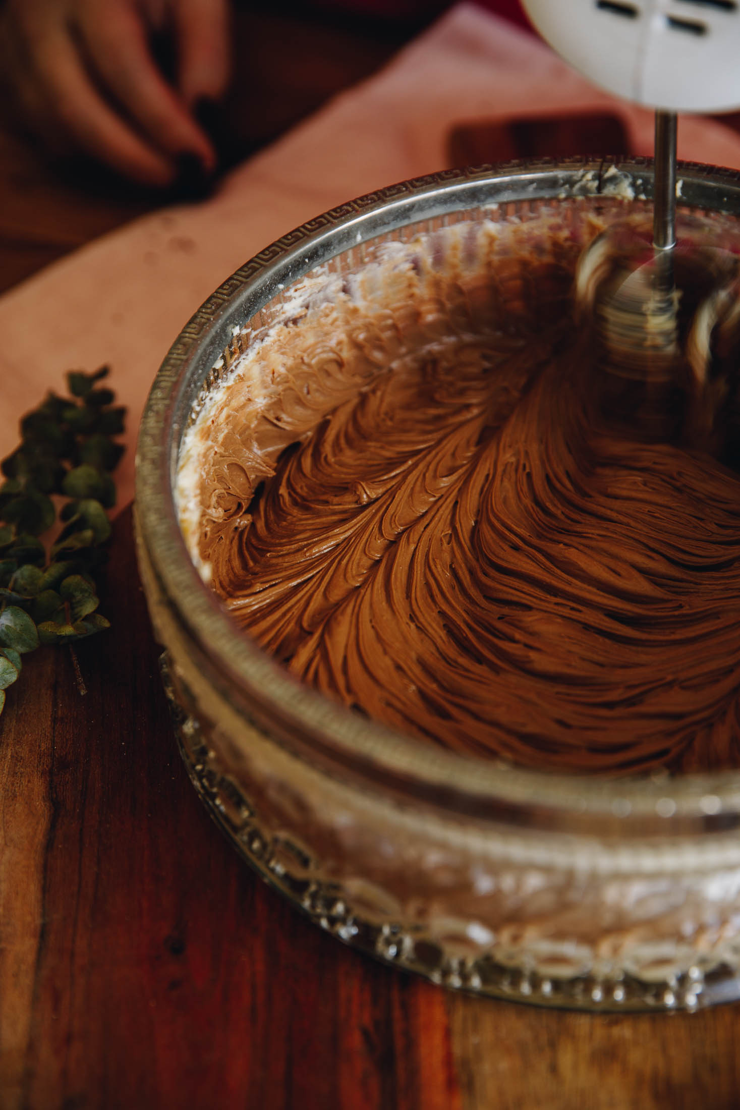 A vintage glass bowl with a silver trim sits on a wooden table. In the bowl is chocolate buttercream that is being whipped with a white hand mixer.