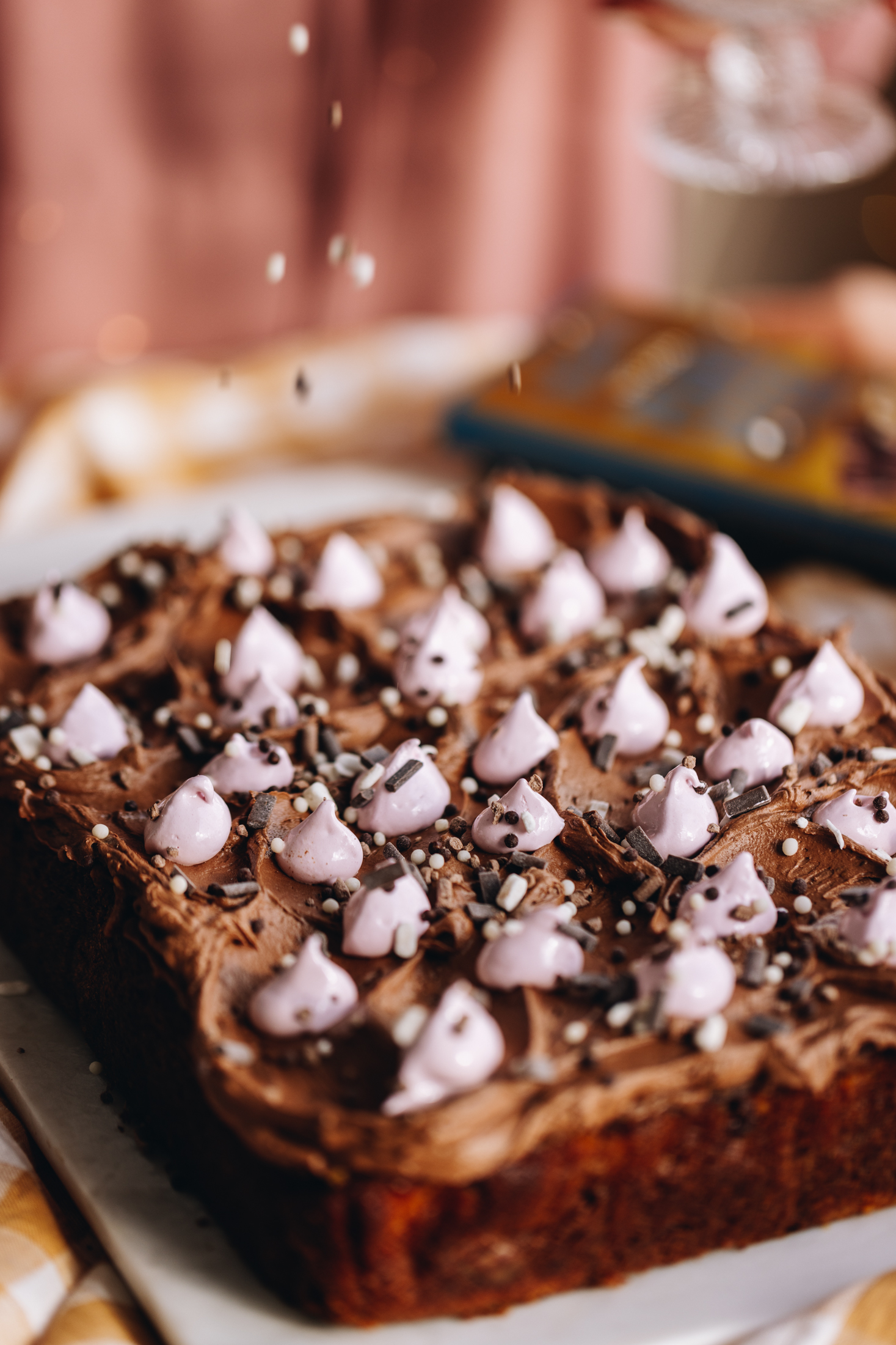 A freshly baked Chocolate peanut butter poke cake is on a white board. The cake has been decorated with a chocolate buttercream and blobs of pink meringue. Brown and white sprinkles are also on it and sprinkles are coming down in the shot.