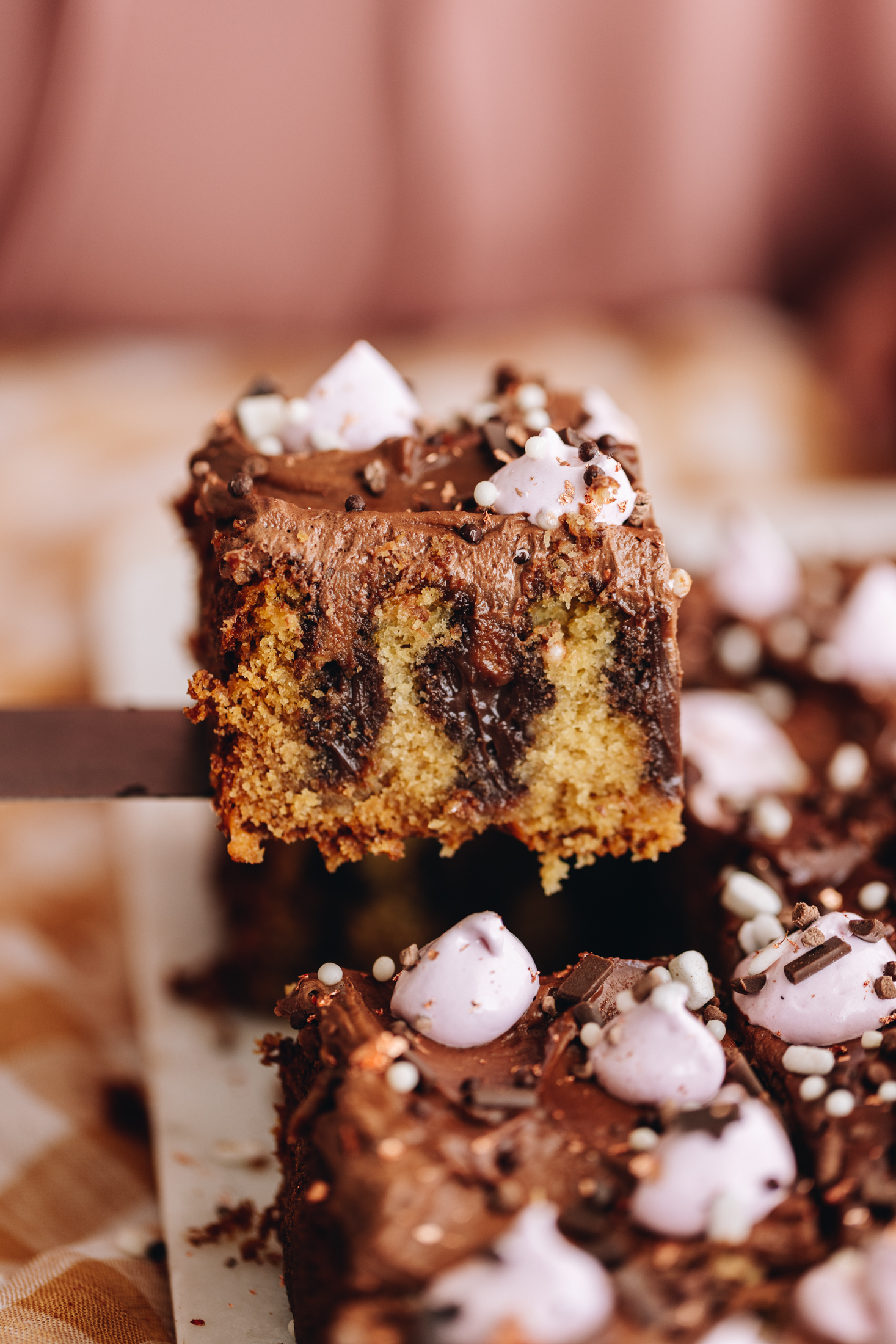 A freshly baked Chocolate peanut butter poke cake is on a white board. The cake has been decorated with a chocolate buttercream and blobs of pink meringue. Brown and white sprinkles are on top. A slice is being help up with a knife. The slice shows the vanilla cake with chocolate ganache poked in to it.