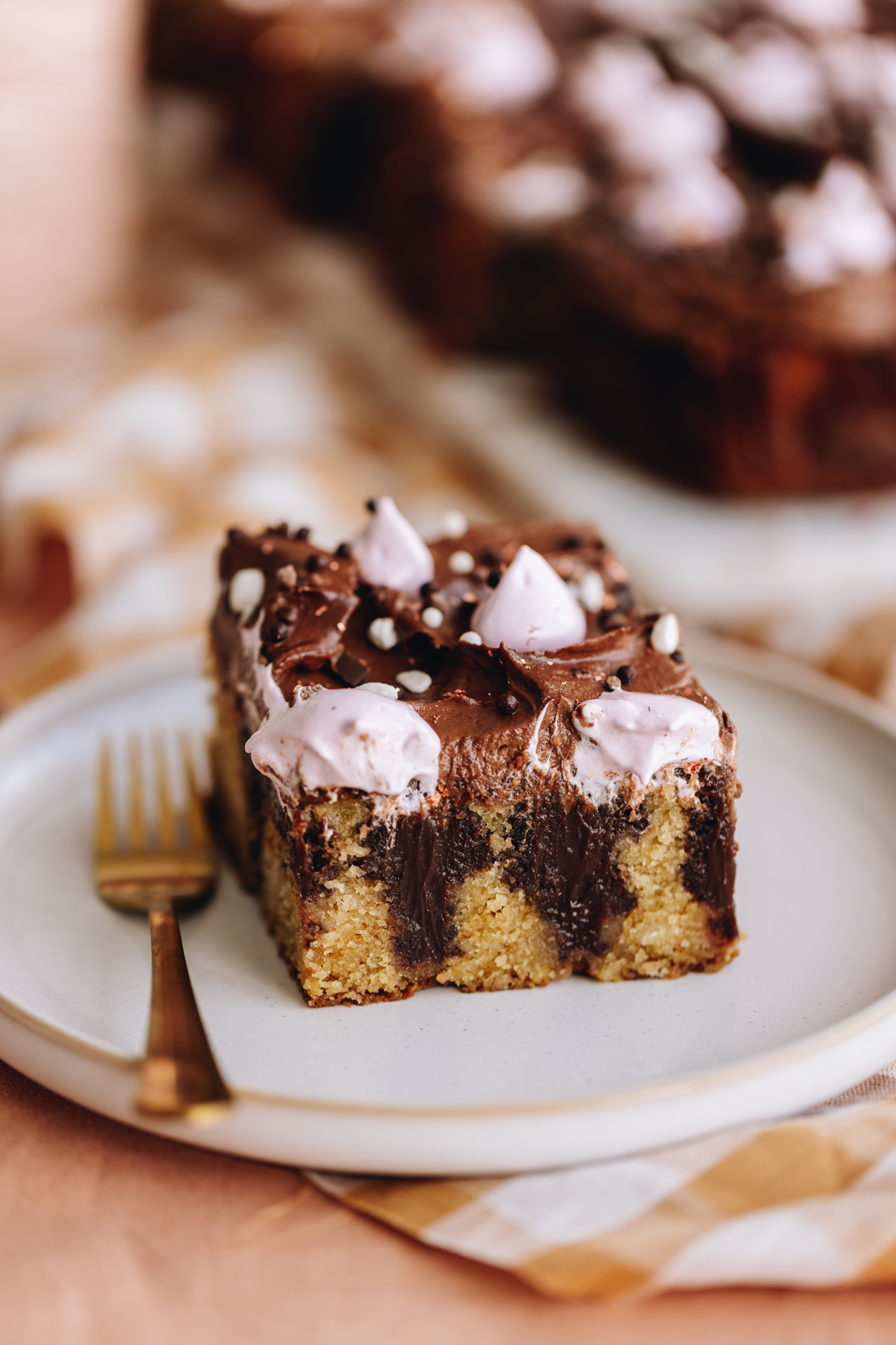 A white plate sits on a pink table cloth with checkered napkins under it. On the plate is a slice of chocolate ganache poke cake. The slice shows the vanilla cake with chocolate ganache poked in to it. It is topped with a chocolate buttercream and pink blobs of meringue. 